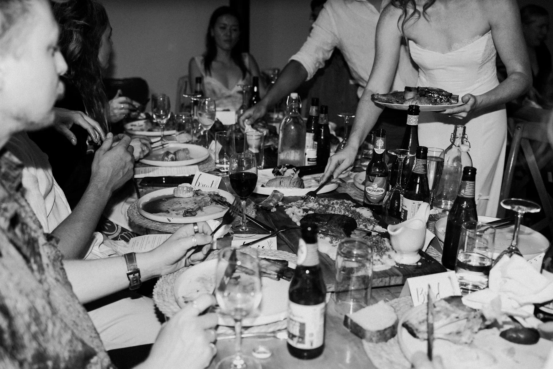 People gathered around a table enjoying a meal, with plates of food, wine glasses, and bottles. A person is serving food from a platter.