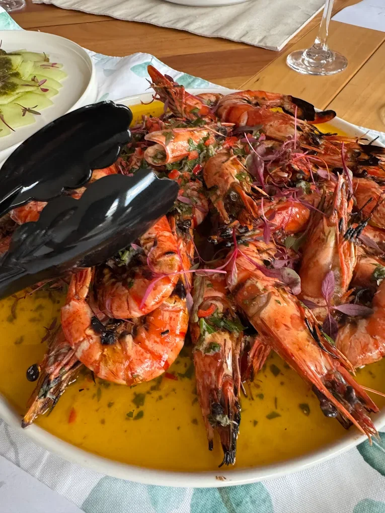 A dish of cooked large shrimp, garnished with herbs, served in a yellow sauce in a white oval platter, on a dining table with some vegetables and a glass of wine nearby.