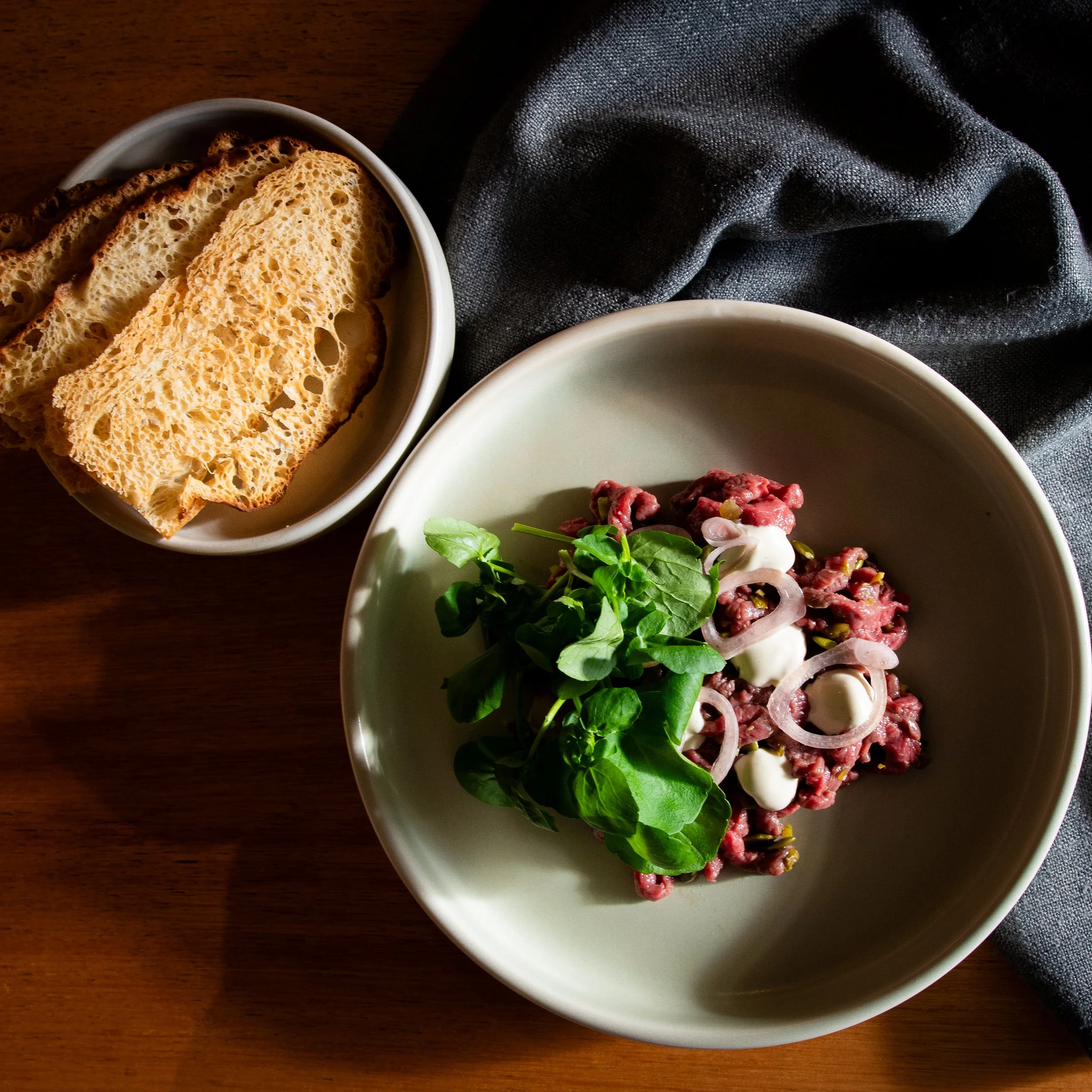 A bowl of sliced toasted bread and a bowl with raw beef, onions, and greens on a wooden table with a dark cloth.