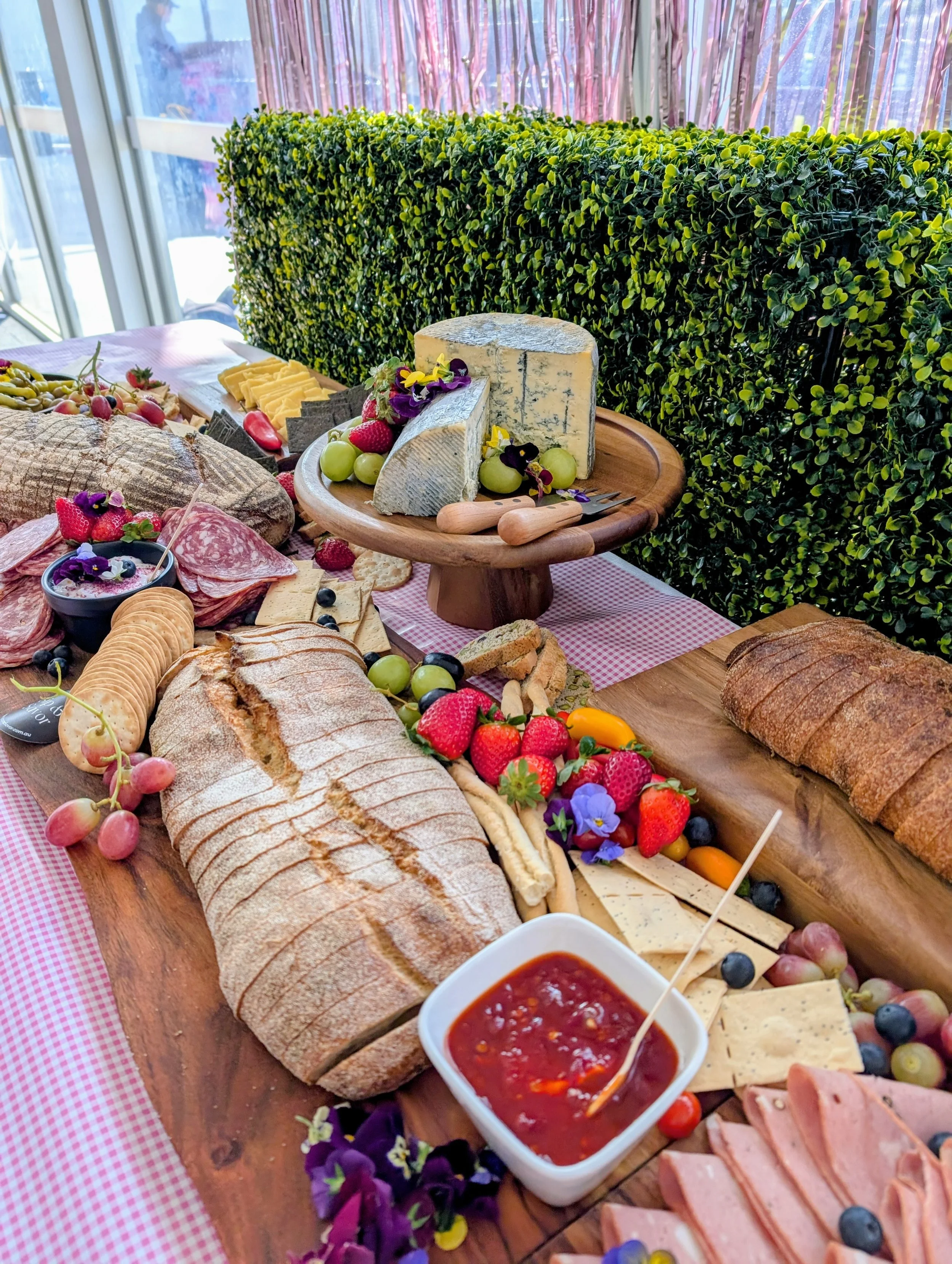 A spread of various cheeses, breads, meats, fruits, and a bowl of jam on a wooden table, with a green hedge and window in the background, and a pink checkered tablecloth.