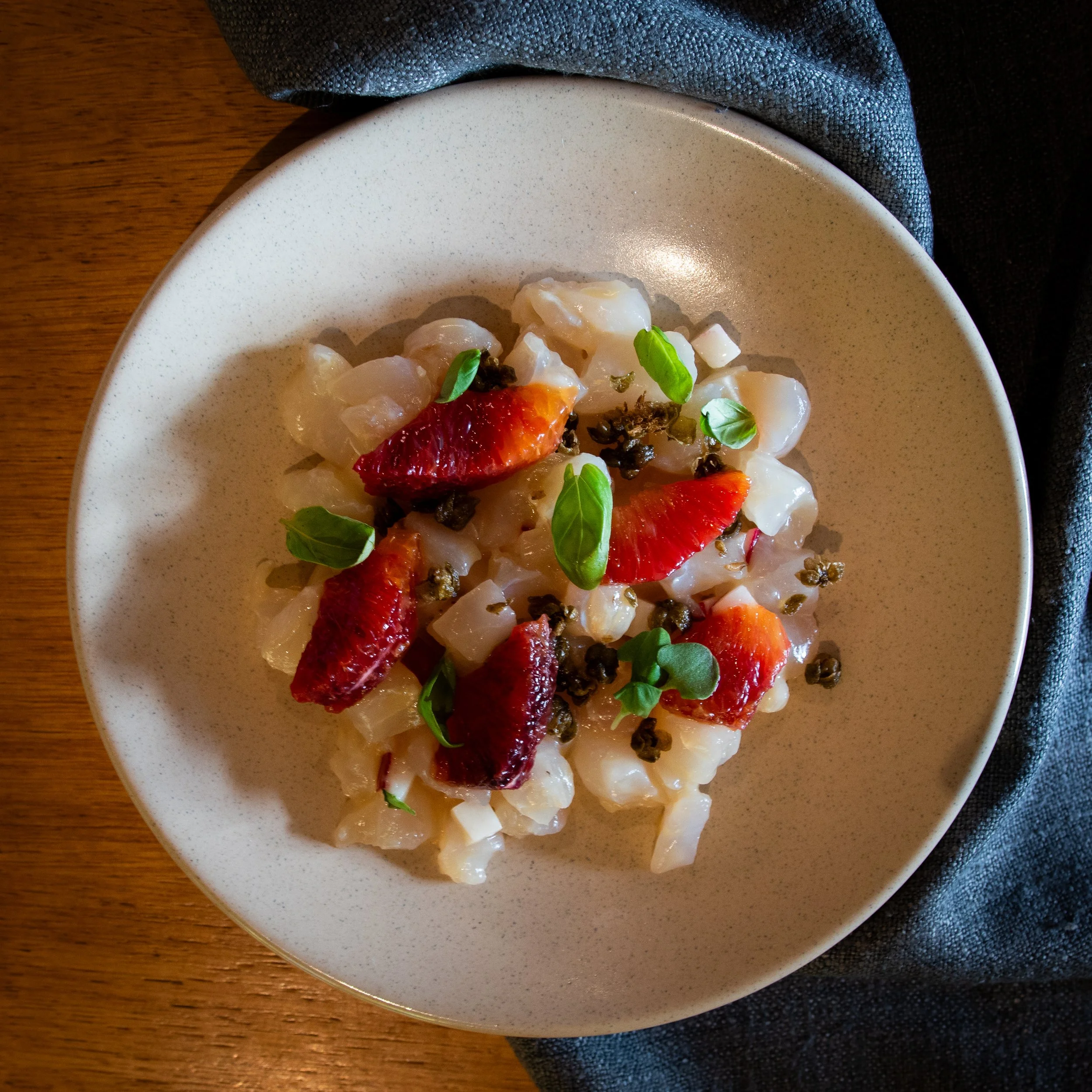 Close-up of a white, speckled ceramic plate with seafood, garnished with fresh green herbs, sitting on a dark placemat on a wooden table.
