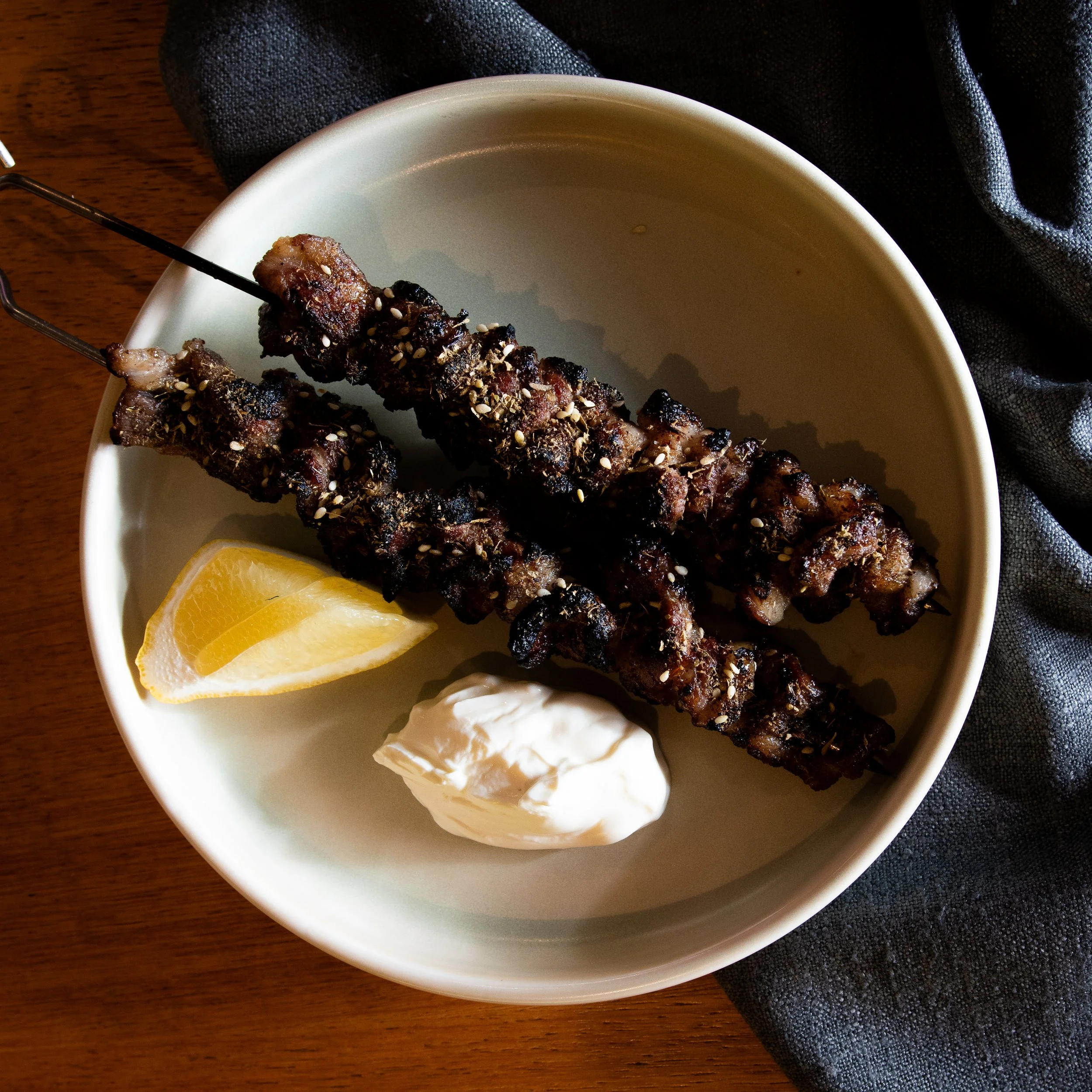Close-up of grilled meat skewers sprinkled with sesame seeds on a white plate with lemon wedges and a dollop of white sauce, on a wooden table with a black napkin.