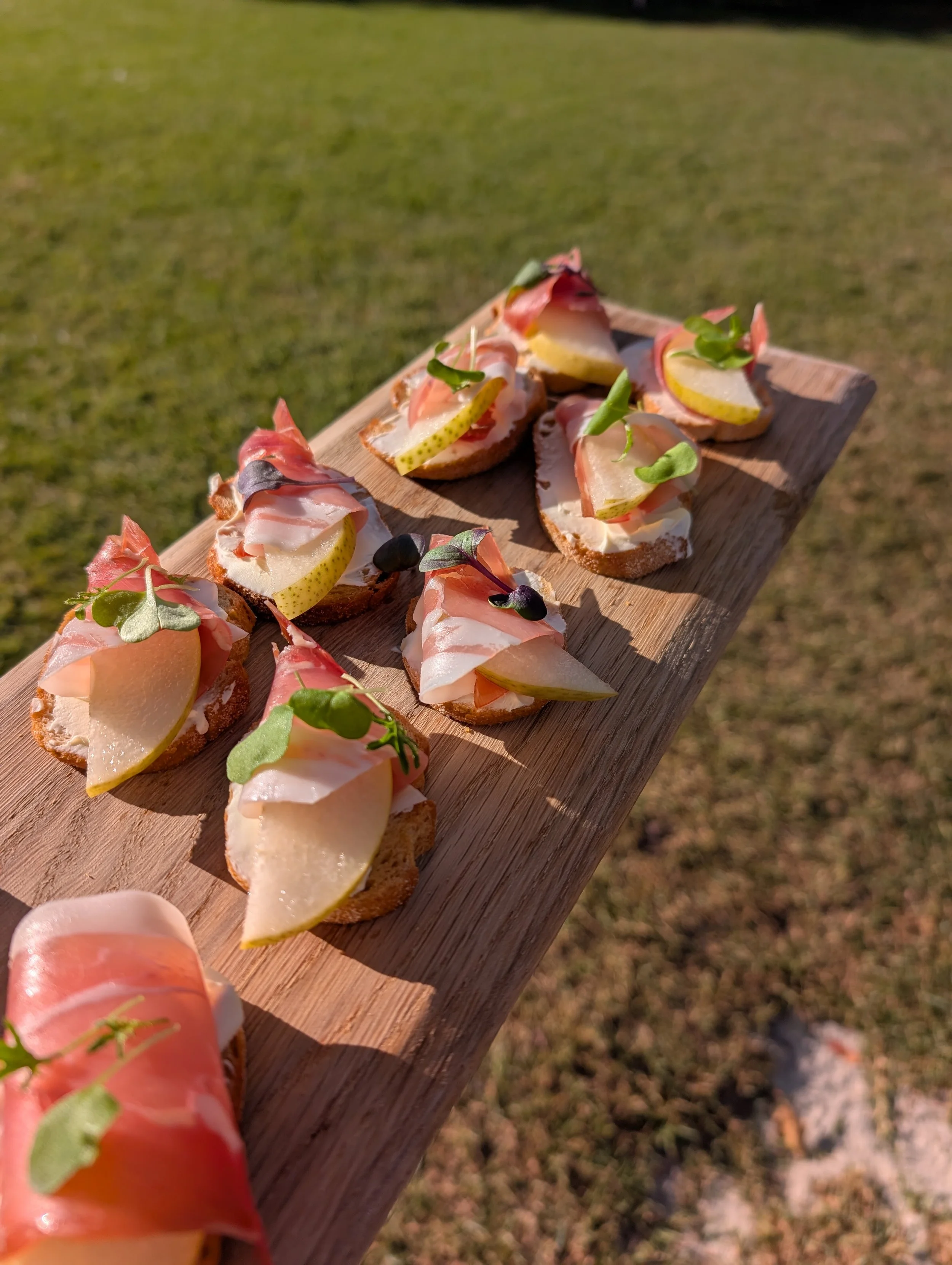 Close-up of small bread slices topped with cheese, thin slices of pear, prosciutto, and garnished with microgreens, on a wooden board outdoors.
