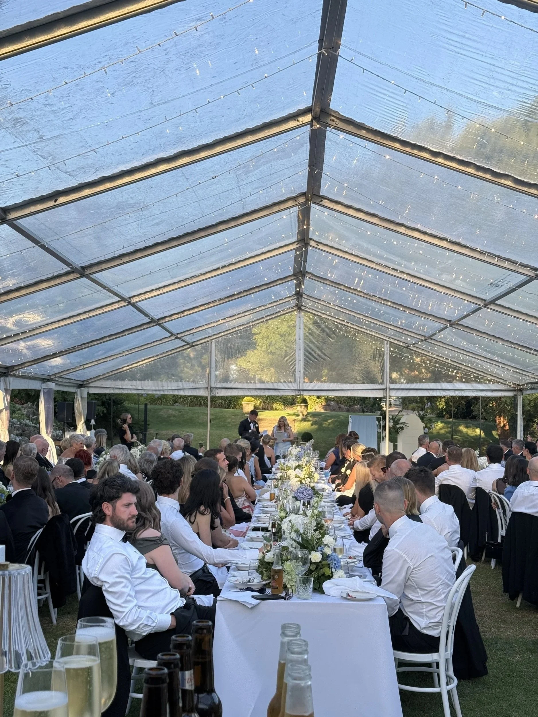 Wedding reception under a tent with guests seated at long tables, a bride and groom giving a speech, and string lights hanging from the tent ceiling.