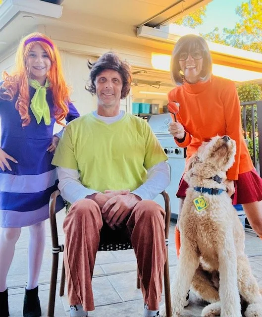 Three people in costumes with a golden retriever dog inside a house. One girl is dressed as a cartoon character with orange hair and a blue dress, standing on the left. A man in a yellow shirt and brown pants is seated in the middle. A woman in an orange dress and glasses is holding a red apple and standing on the right, with the dog sitting and looking up.