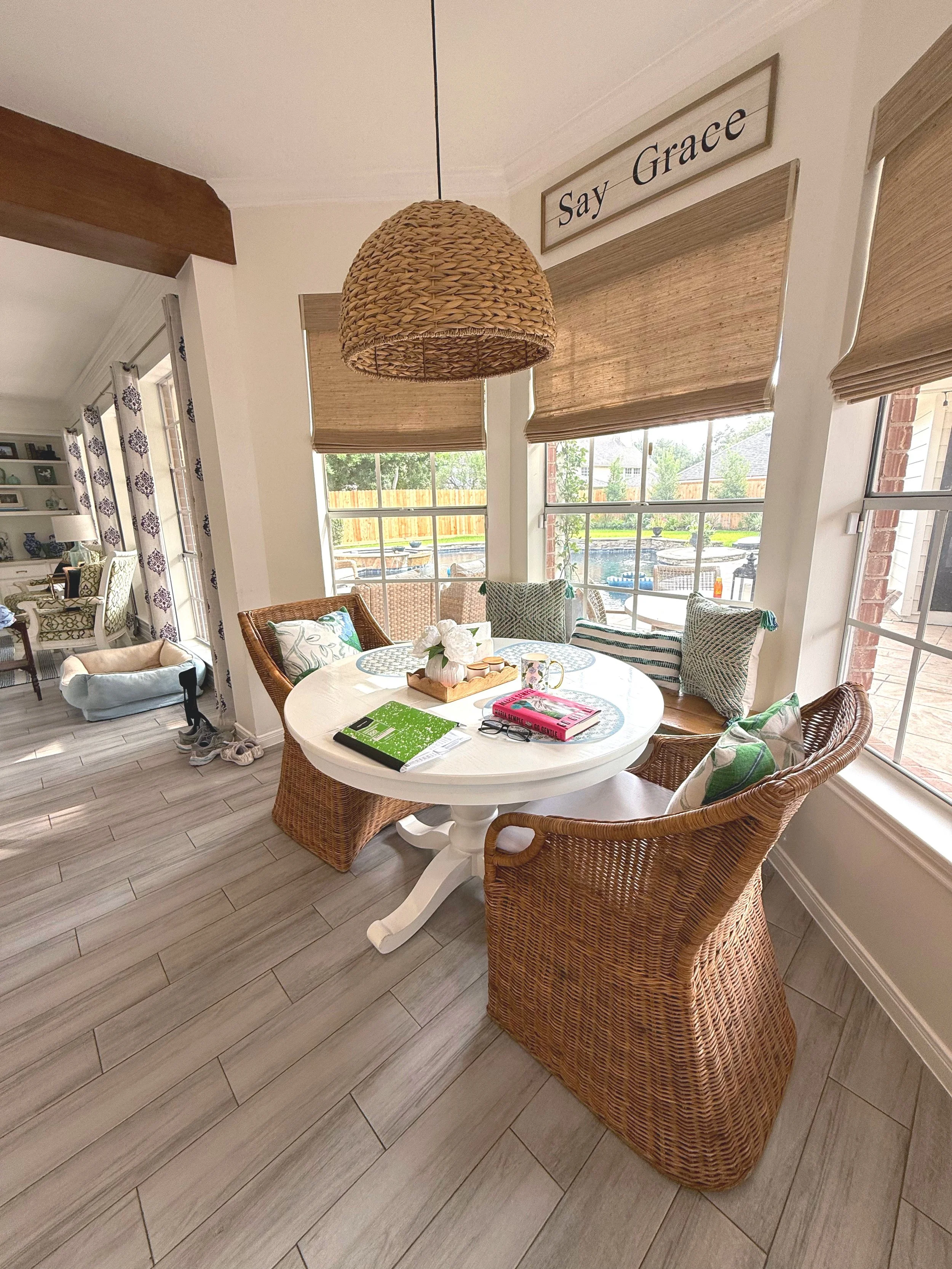A bright dining area with a round white table surrounded by wicker chairs, with a window view of a backyard pool and outdoor furniture, and a sign saying 'Say Grace' on the wall.