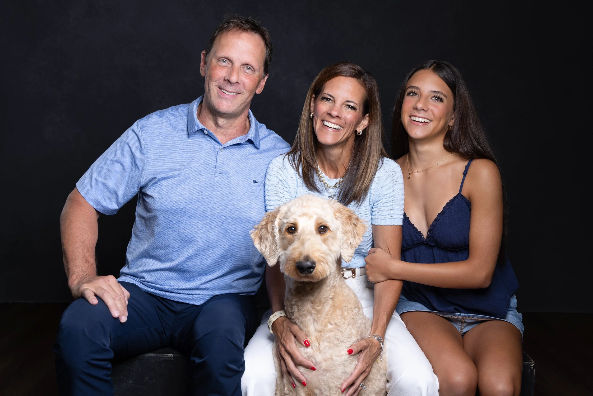 A smiling family of three with a golden retriever puppy against a black backdrop. The father is wearing a blue polo shirt, the mother is in a light blue top, and the daughter is in a dark blue dress. The mother is holding the puppy, and everyone is smiling.