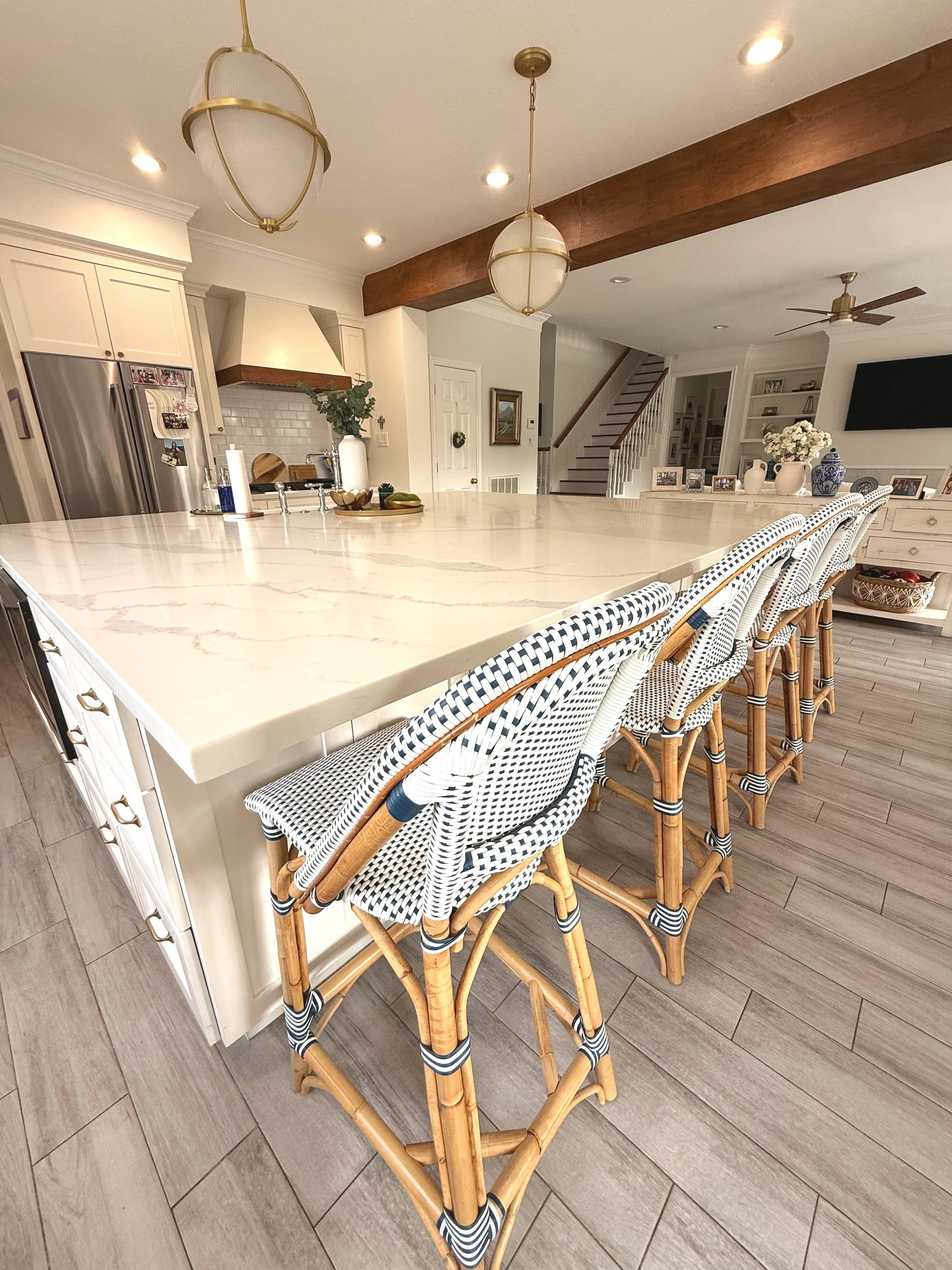 A kitchen with a white island counter, wicker bar stools with black and white striped cushions, stainless steel refrigerator, white cabinets, a wooden beam ceiling detail, hanging pendant lights, a staircase with wooden steps and white railing in the background, and a wall with framed photos and decorations.