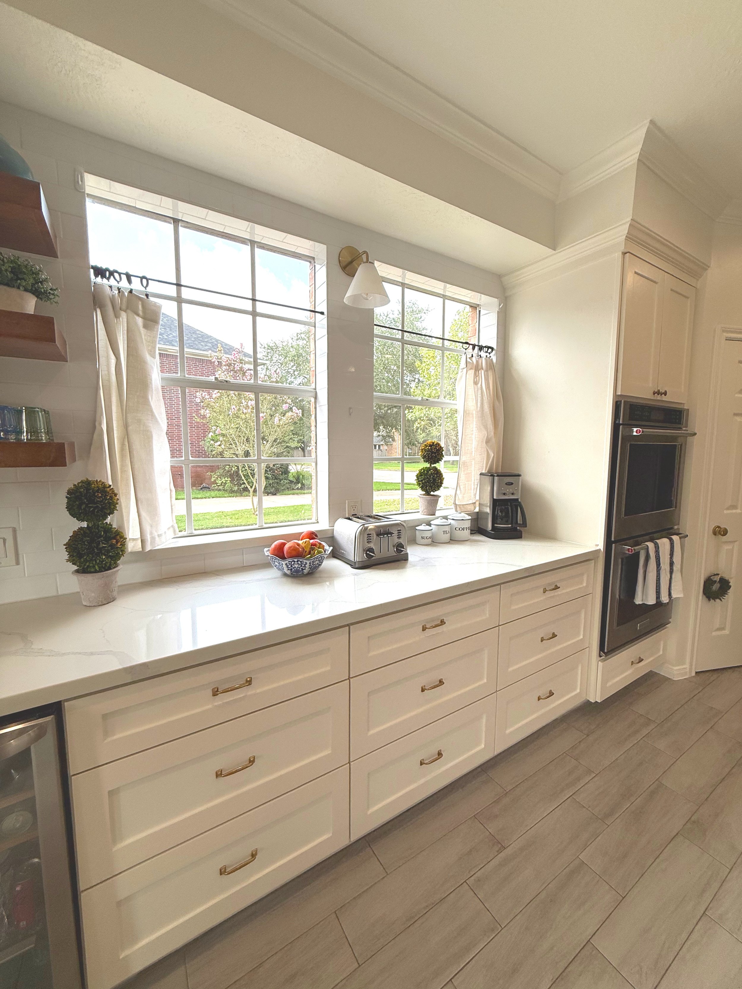 Bright kitchen with large window, white cabinets with gold handles, and a marble countertop. On the counter are a fruit bowl, toaster, coffee maker, and labeled containers for sugar, tea, and coffee. There are decorative plants and shelves on the wall.