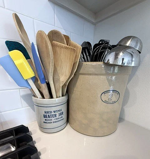 Kitchen countertop with wooden spoons and utensils in a ceramic holder, and metal measuring cups in a ceramic container.