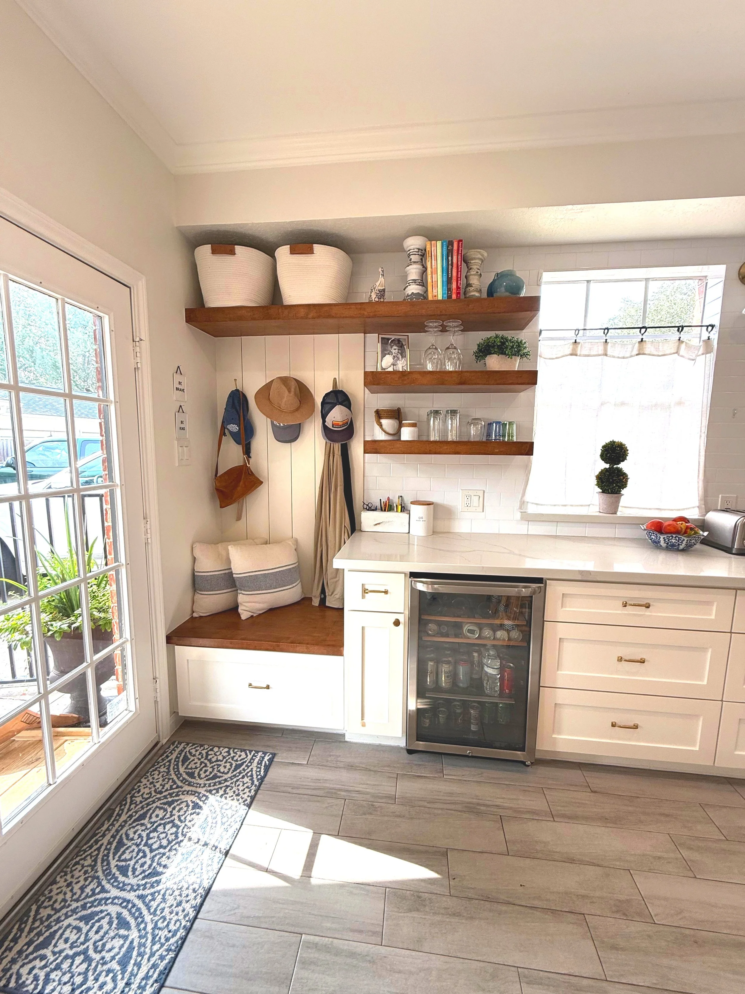 Coffee station in a bright kitchen with white cabinetry, open wooden shelves with books, decorative vases, glasses, and potted plants, a small wine cooler, and a window with a light curtain.
