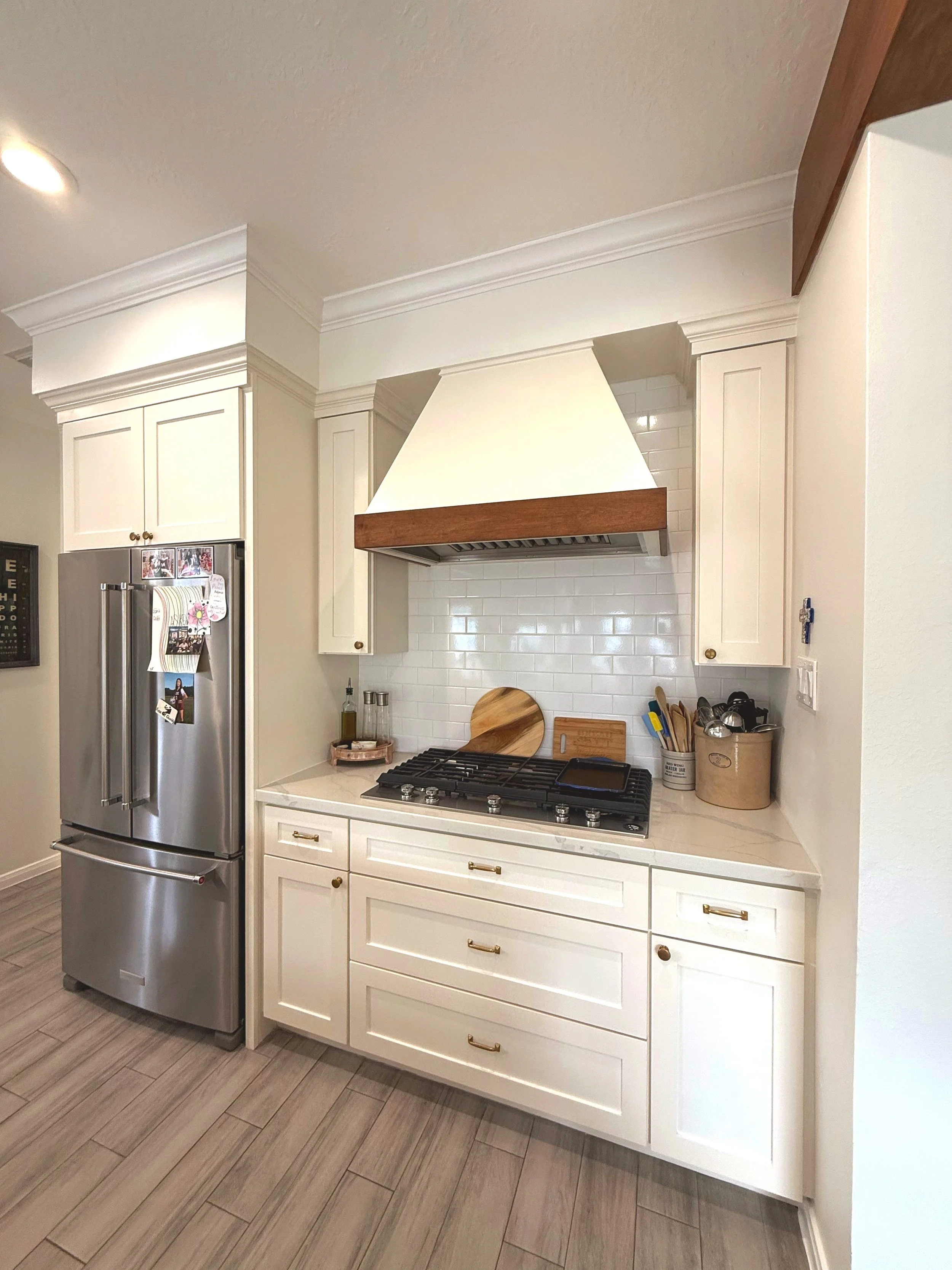 Modern kitchen with white cabinets, a stainless-steel refrigerator, a gas stove, and a white range hood with wood trim. The cabinetry has gold handles, and the countertops are white. The floor is wood-look tile.