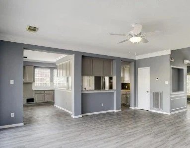Empty living room with gray walls, white ceiling fan, wood flooring, and large windows