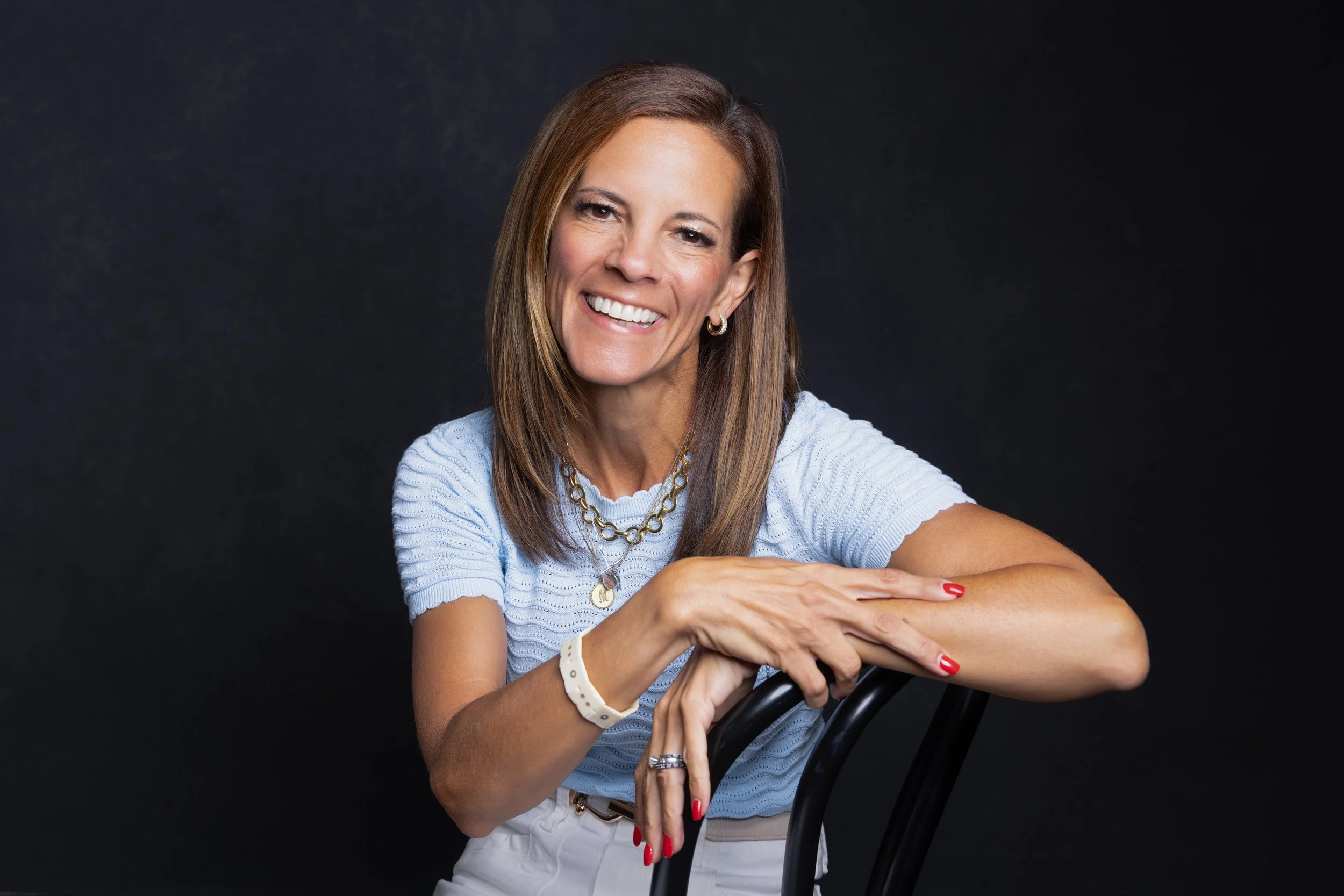 A woman with long brown hair, smiling, wearing a light blue top and jewelry, sitting on a chair against a dark background.