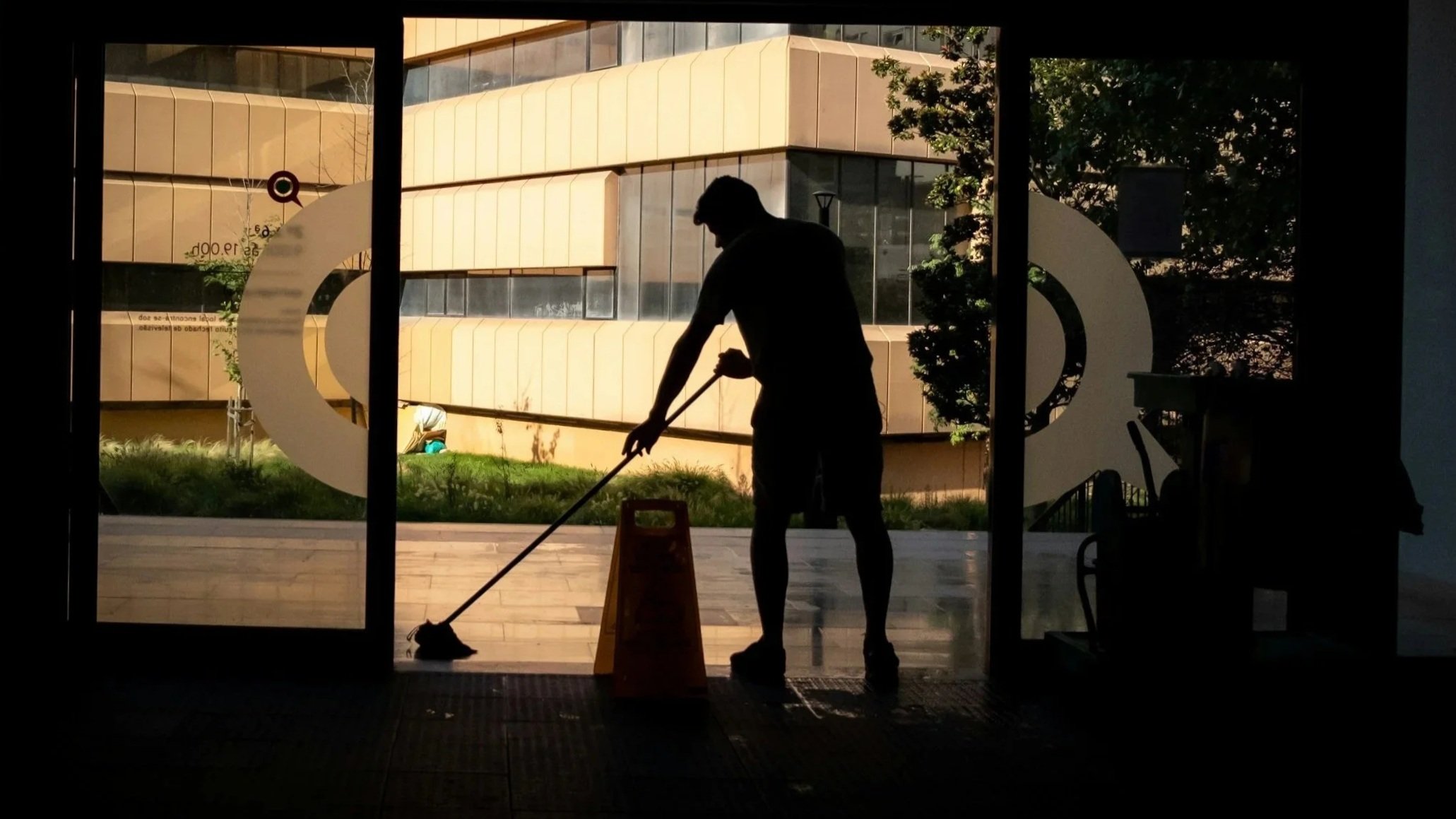 Silhouette of a person cleaning the floor inside a building, seen through glass doors with an external view of a modern building and trees.