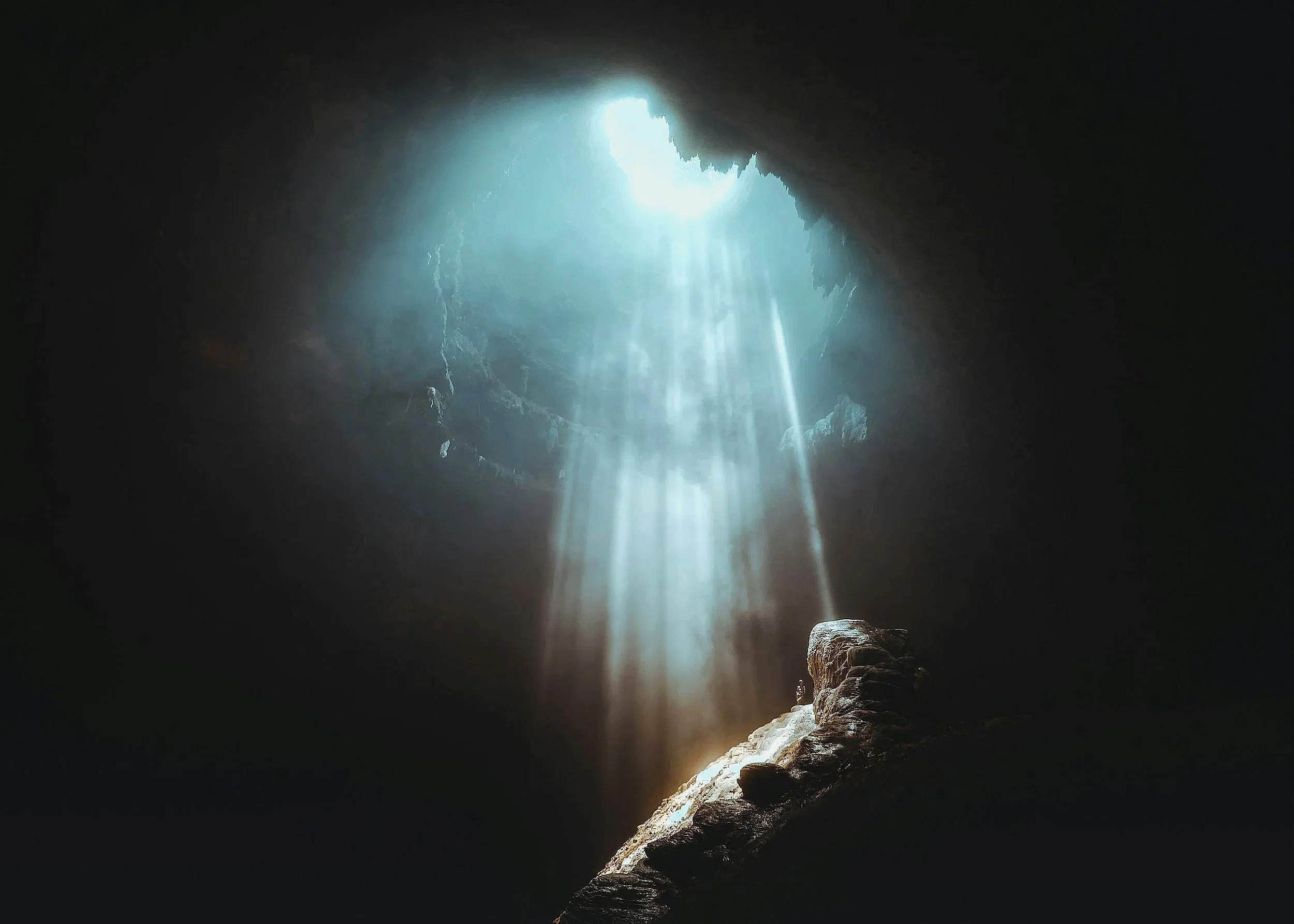 Sunlight streams into a dark cave through an opening at the top, illuminating a rocky interior and a small pile of stones on the cave floor.