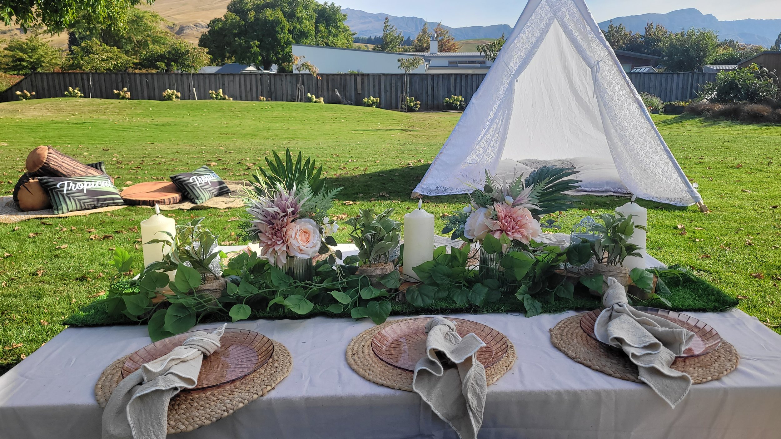 Outdoor setting with a white canopy, floral decorations, candles, and place settings on a table in a grassy yard with trees and mountains in the background.