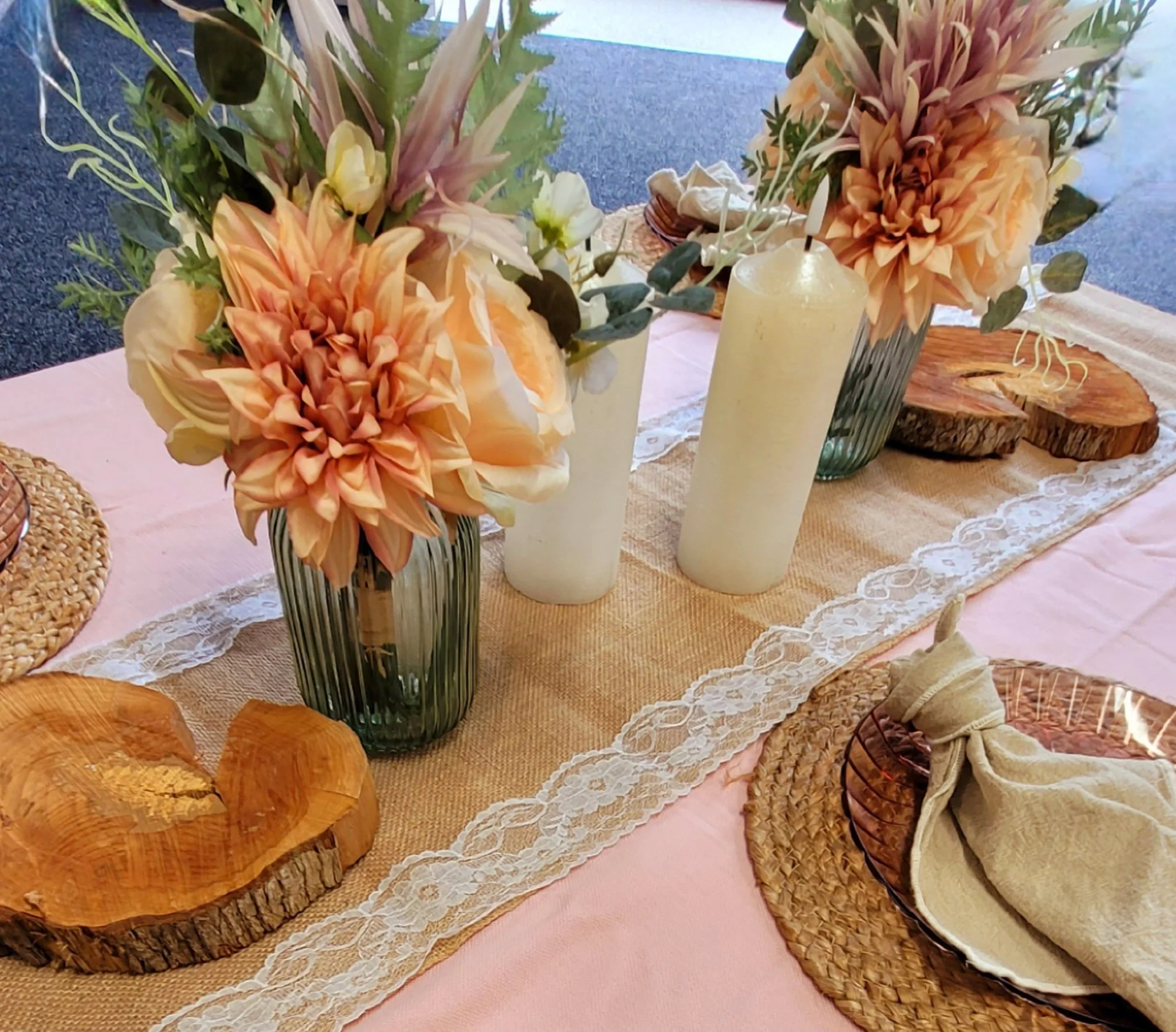 Floral centerpiece with large peach and cream-colored flowers in a striped glass vase on a wooden table with rustic wood slices and a candle, decorated with lace, on a pink tablecloth.