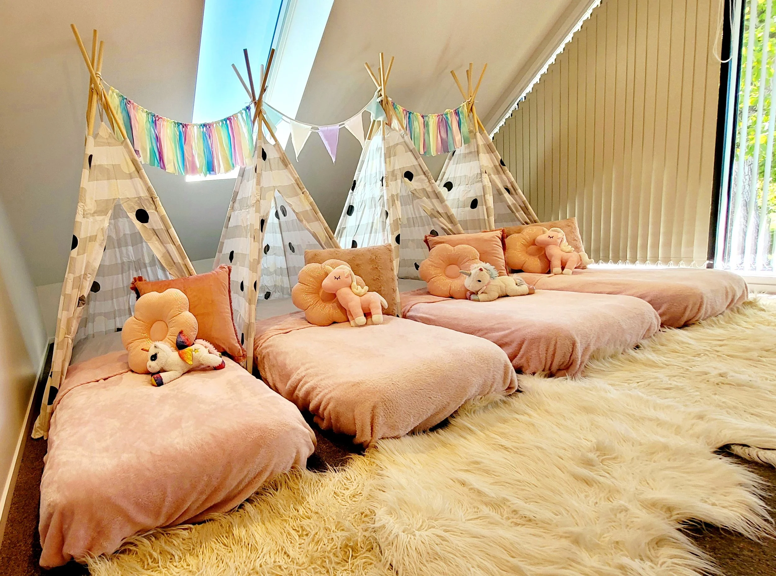 A cozy children's play area with pink plush seating, stuffed unicorn and cloud toys, teepees with polka dot fabric, colorful bunting, and a large window with vertical blinds, all on a fluffy cream rug.