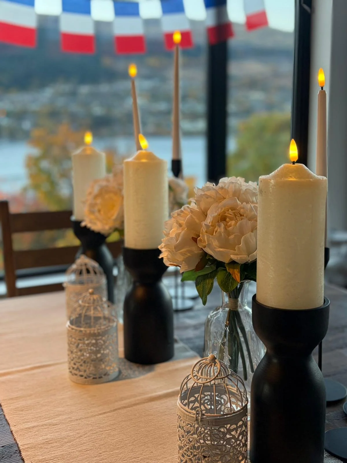 Decorative table centerpiece featuring white candles in black holders, white floral arrangements in glass vases, and white ornamental birdcages, with a background of a large window and a blurred view outside.