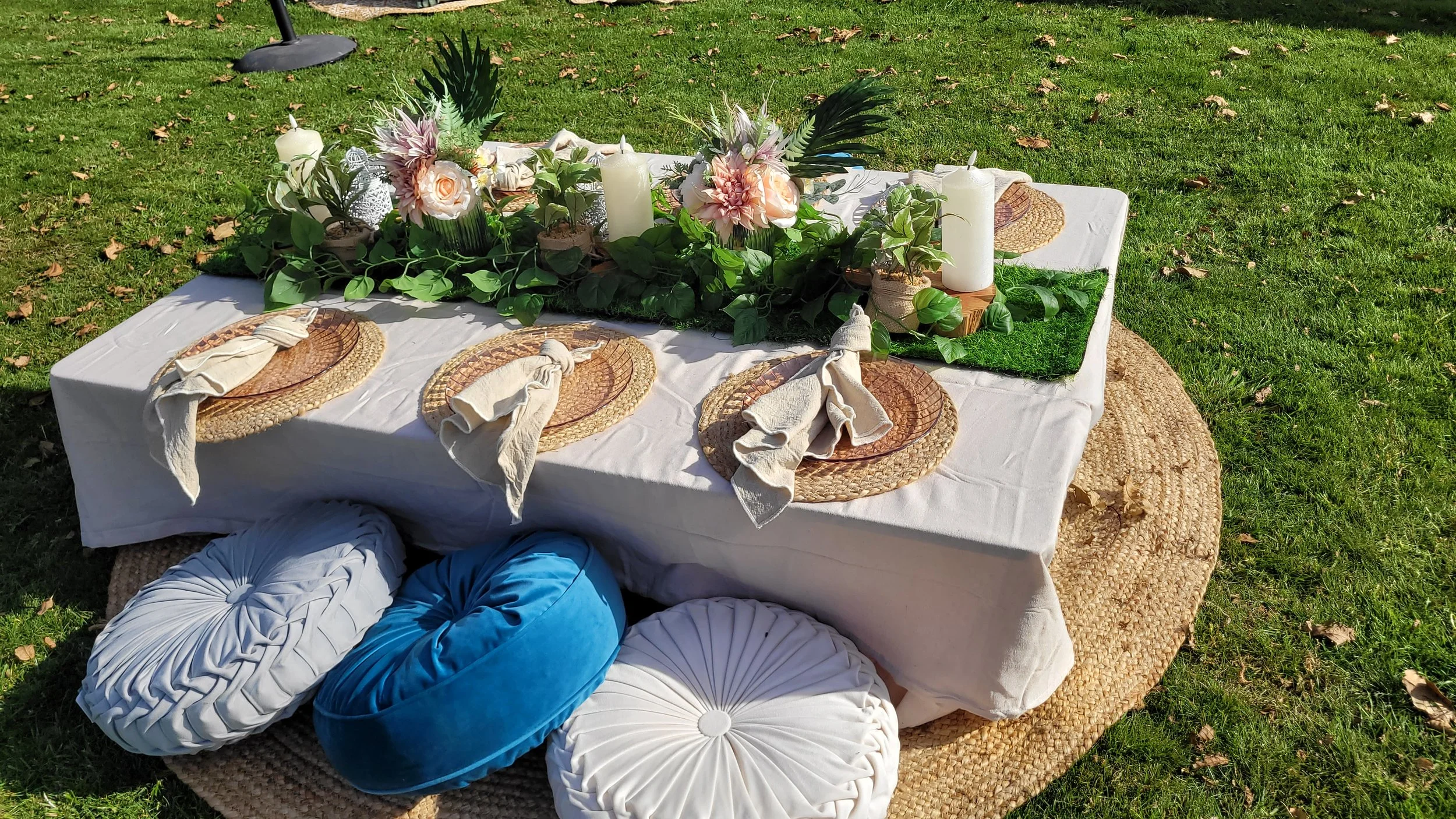 Outdoor table setting with a white tablecloth, woven placemats, beige napkins, and pillows in shades of white and blue, surrounded by green grass, featuring a floral centerpiece with white candles, pink and peach flowers, and green foliage.