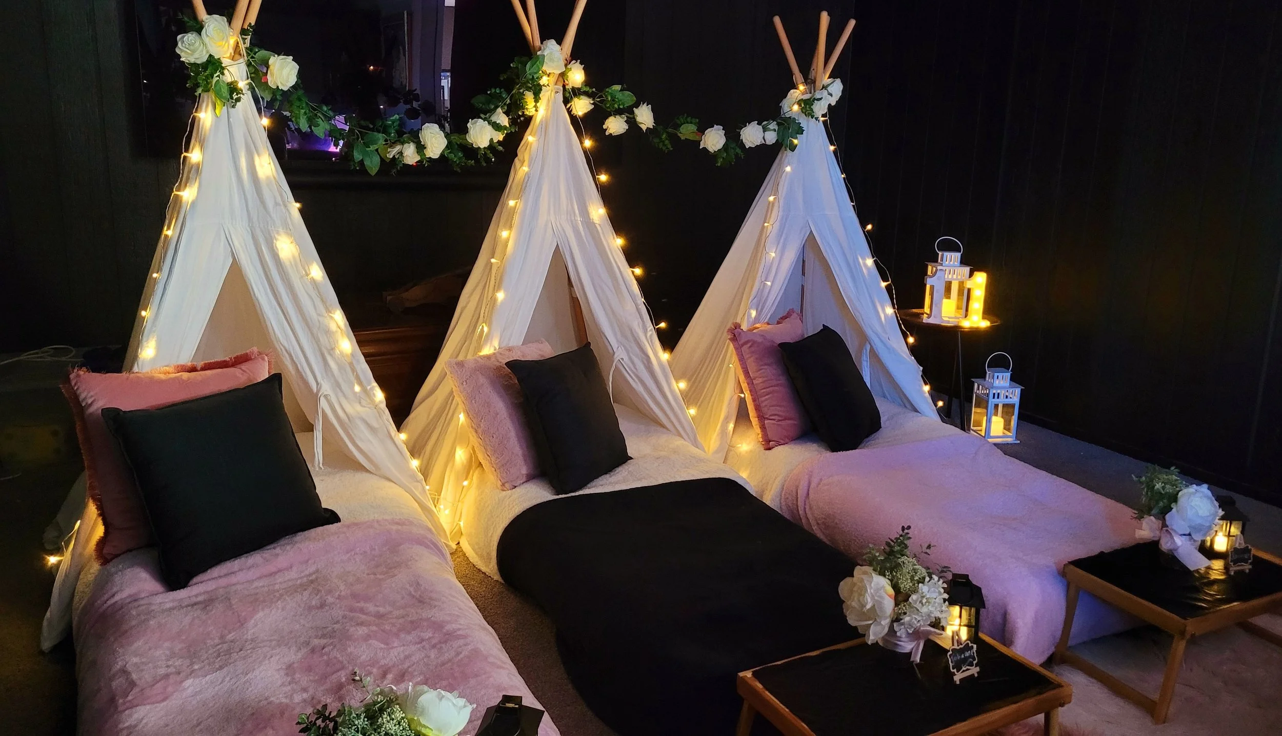 Decorative indoor setup with three small white fabric tents illuminated by string lights, each tent decorated with floral garlands and pillows, placed on a pink bedding-covered surface. Nearby, there are lanterns and floral arrangements, creating a cozy, festive atmosphere.