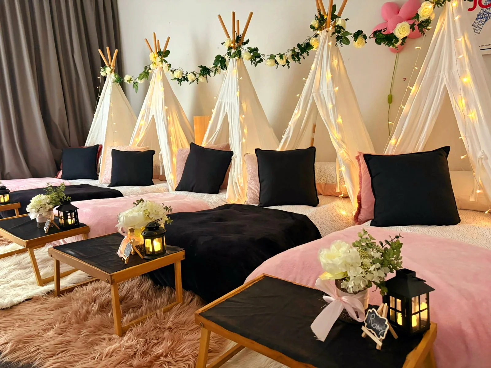 Decorated room with white teepees draped with string lights, black pillows, pink bedsheets, plants, and small tables with lanterns and flowers.