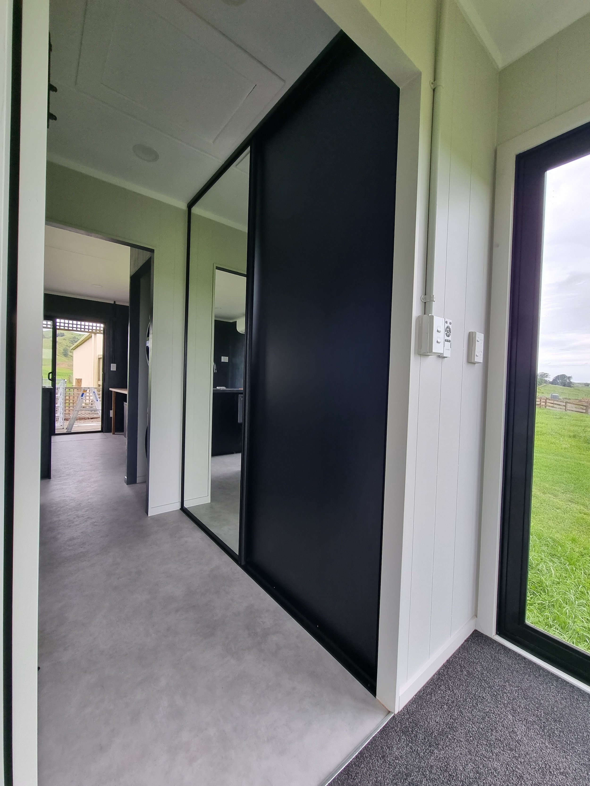 Interior of a modern house with a hallway, black sliding door, white walls, large window showing green field outside, and bathroom in background.