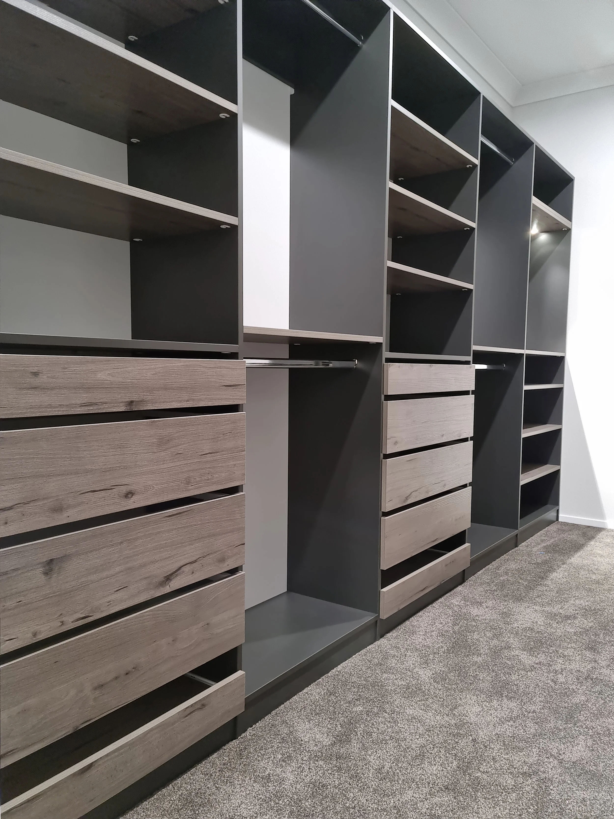 A modern storage closet with multiple open shelves and drawers made of wood and dark gray panels, situated in a room with a gray carpeted floor and white walls.