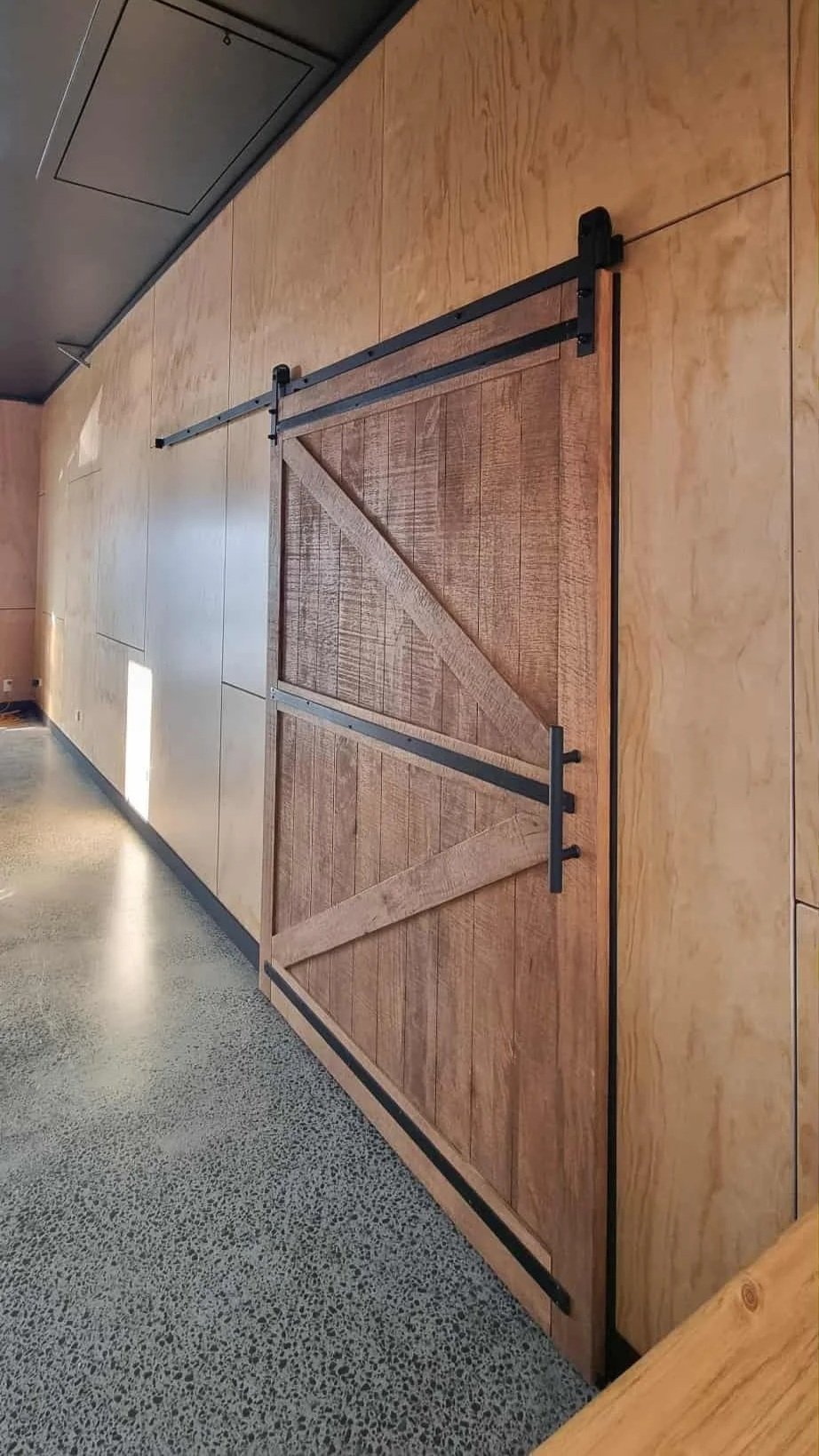A wooden sliding barn door with black metal hardware on a light wood-paneled wall.