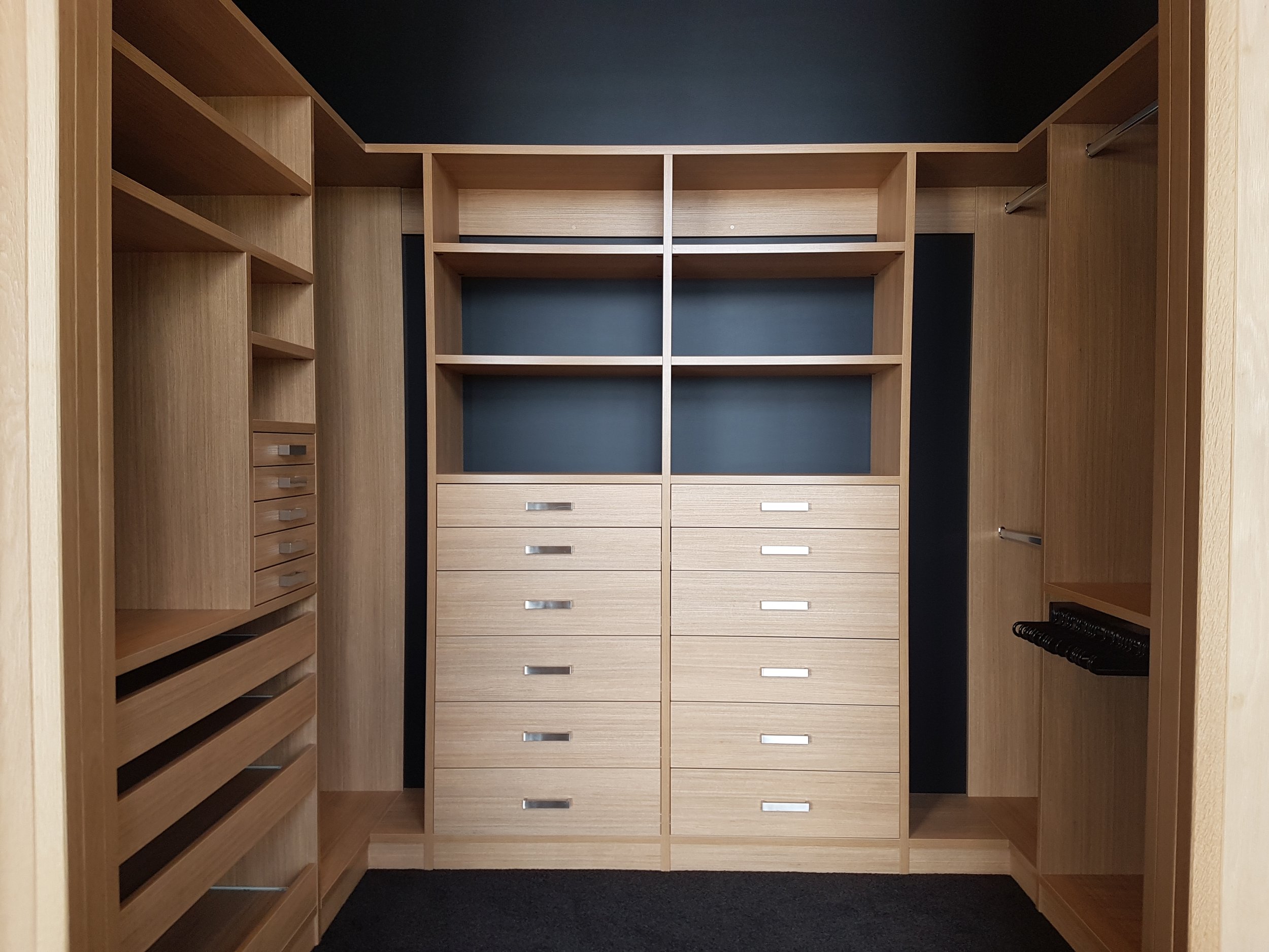 A walk-in closet with wooden shelves, drawers, and hanging rods, with dark walls and a black carpeted floor.