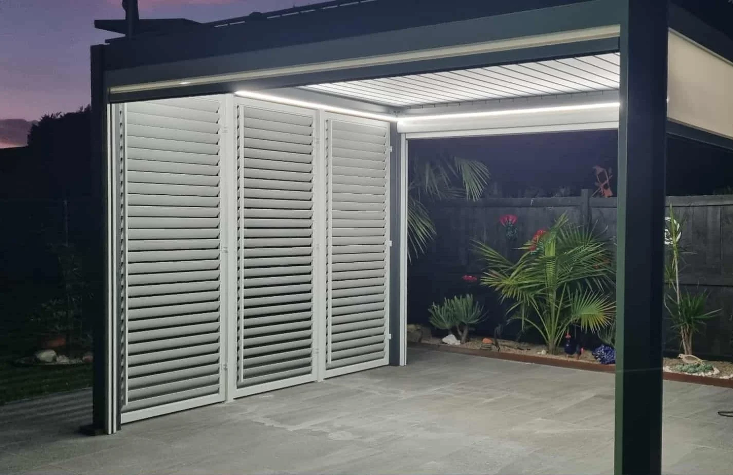 A modern outdoor patio area with a metal pergola featuring an integrated LED light strip, white louvered shutters, and a garden with various green plants and decorative garden ornaments.