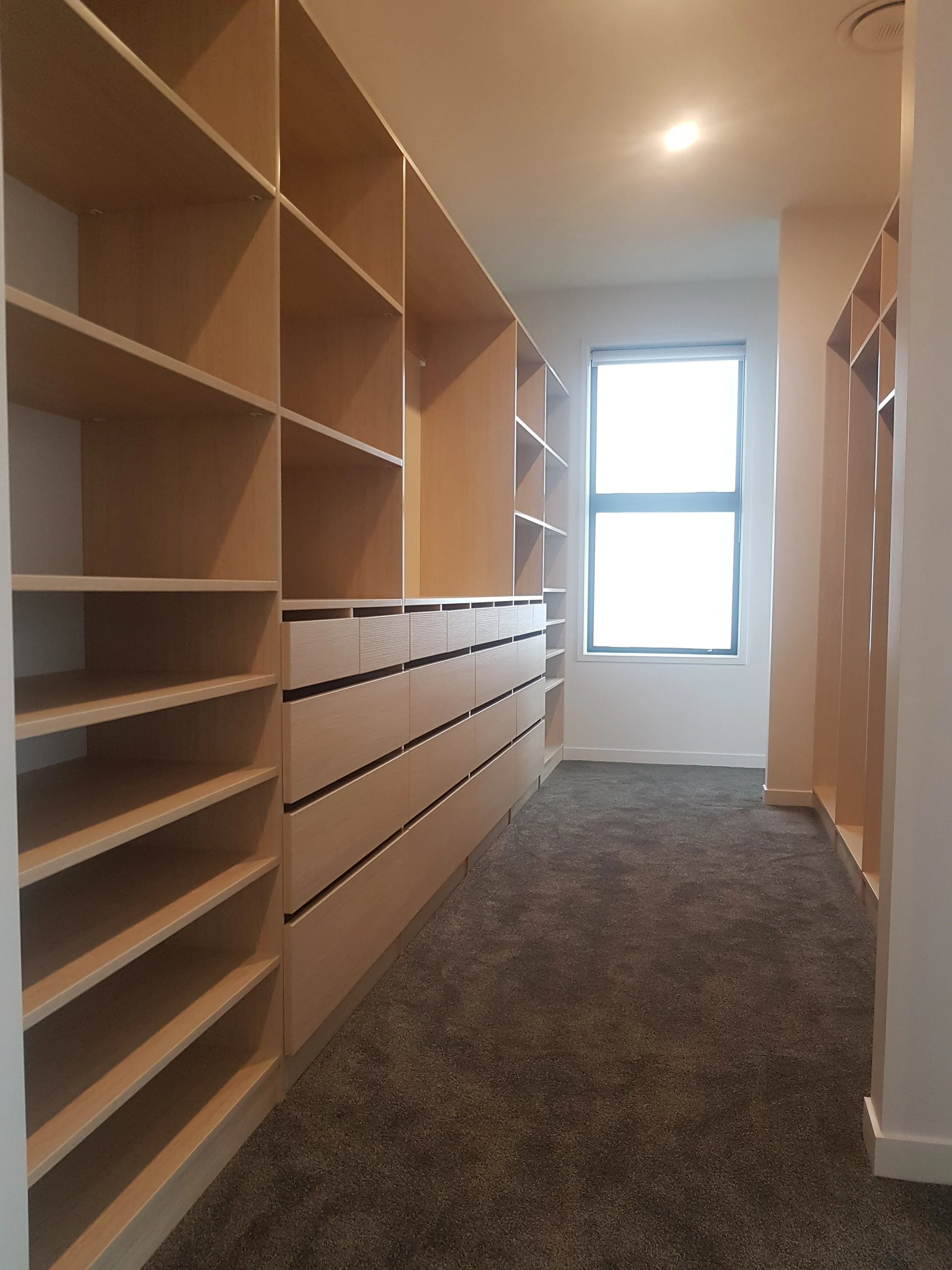 A walk-in closet with wooden shelves and drawers, and a window at the end allowing natural light.