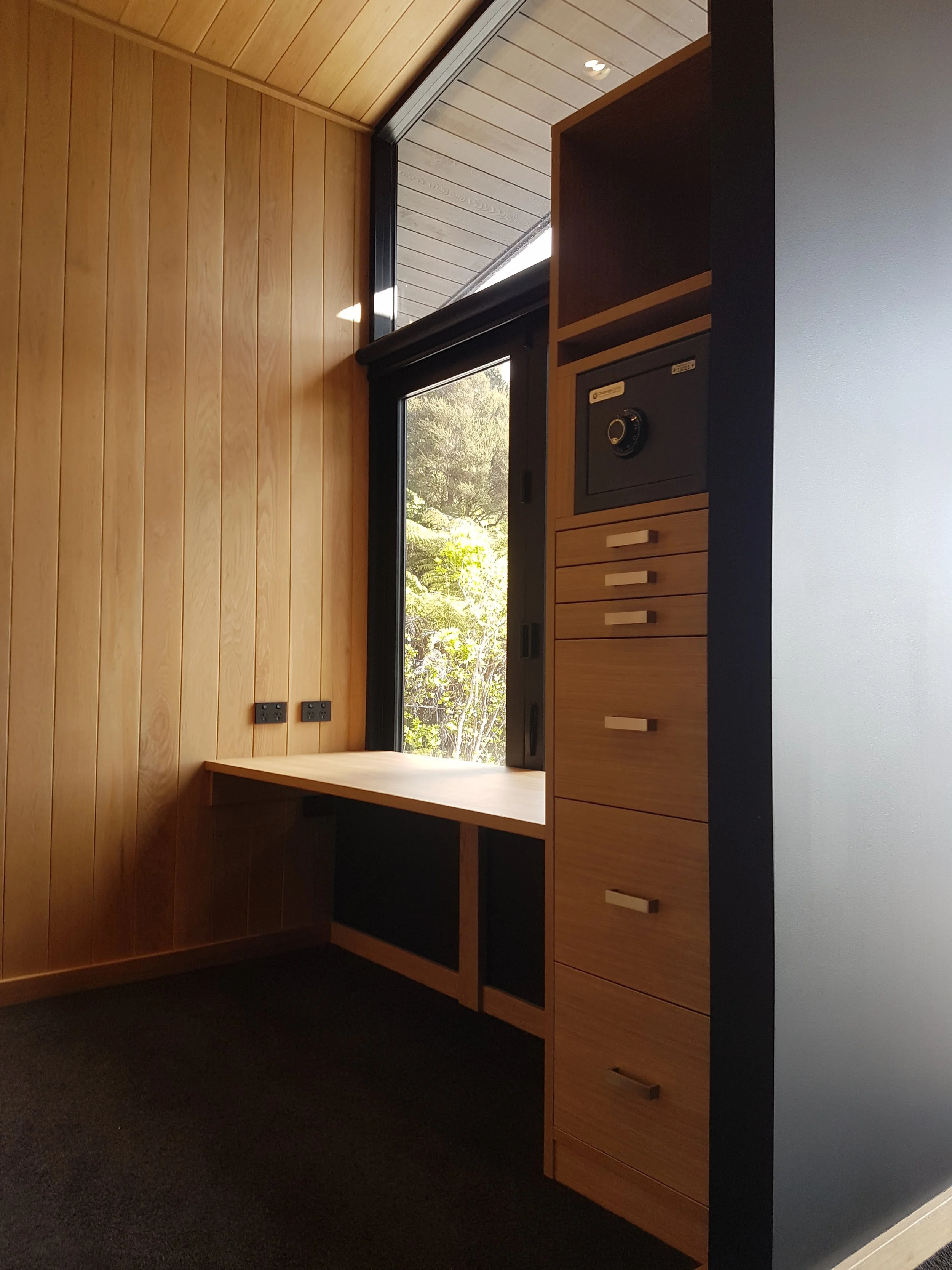 A room with wooden panel walls and a black countertop with wooden drawers. There is a window showing a view of trees and bushes outside.