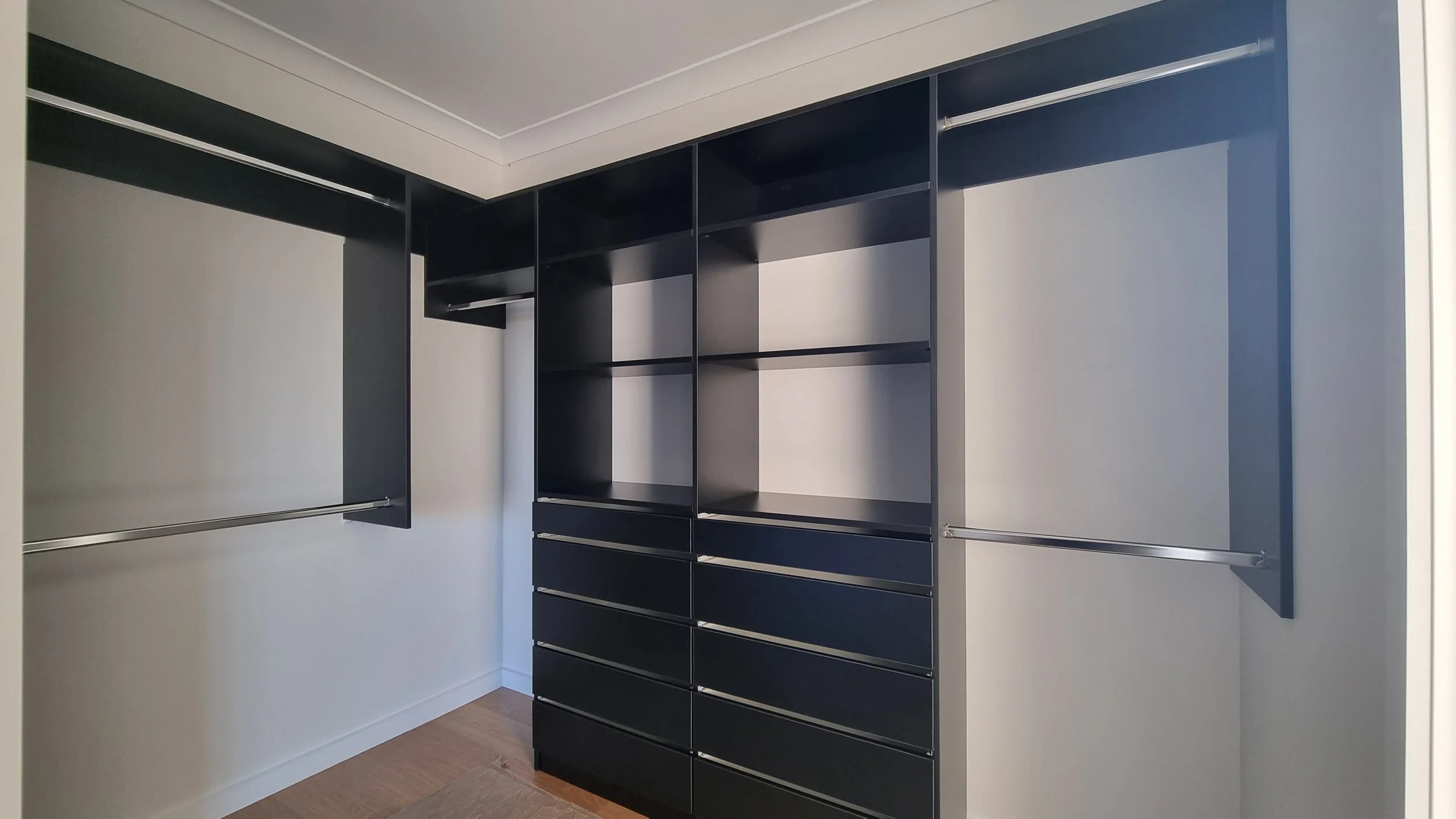 Empty walk-in closet with black shelving and hanging rods, white walls, and hardwood floor.