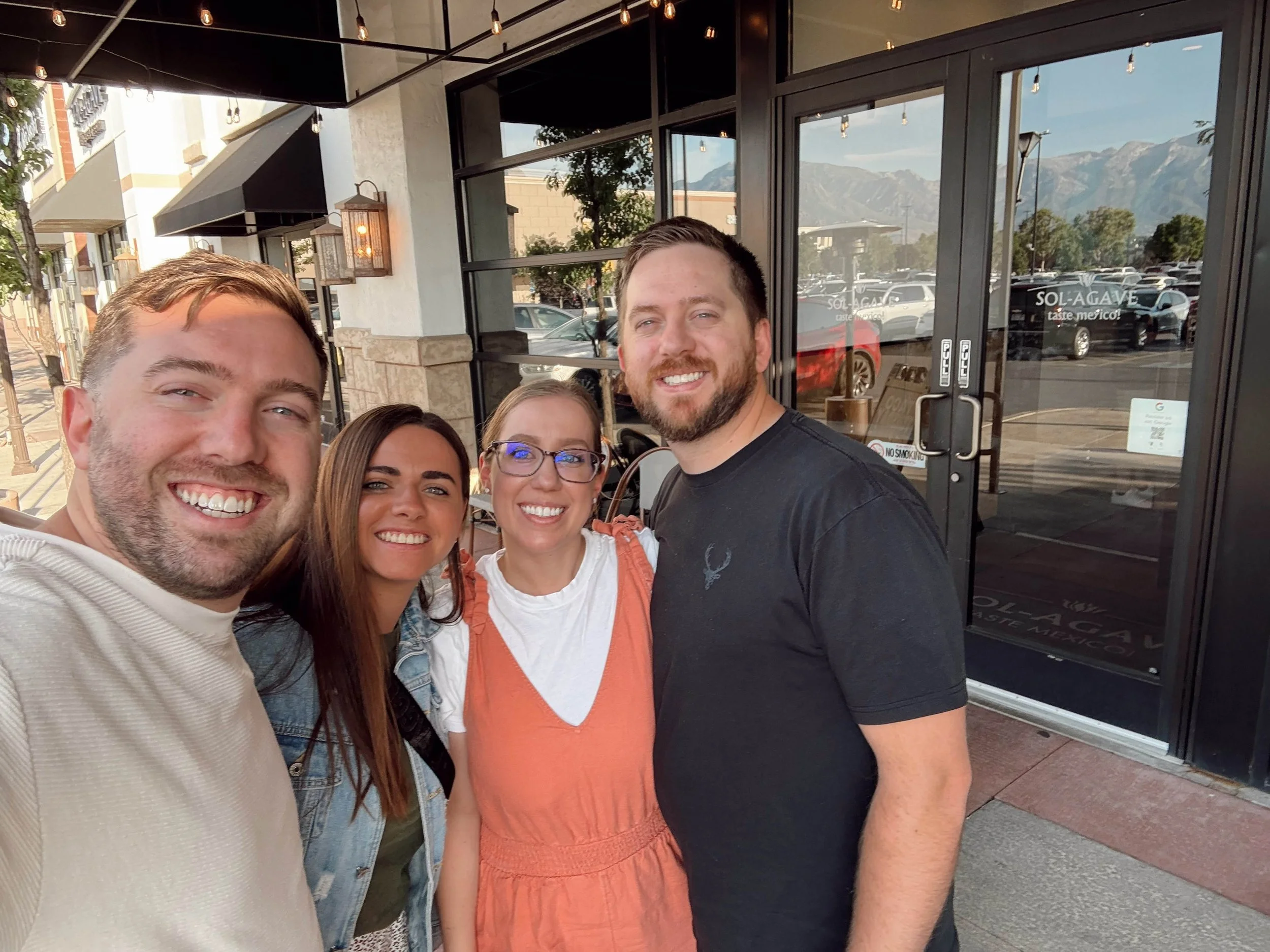Group of four people taking a selfie outside a restaurant or cafe, with glass doors and mountains in the background.