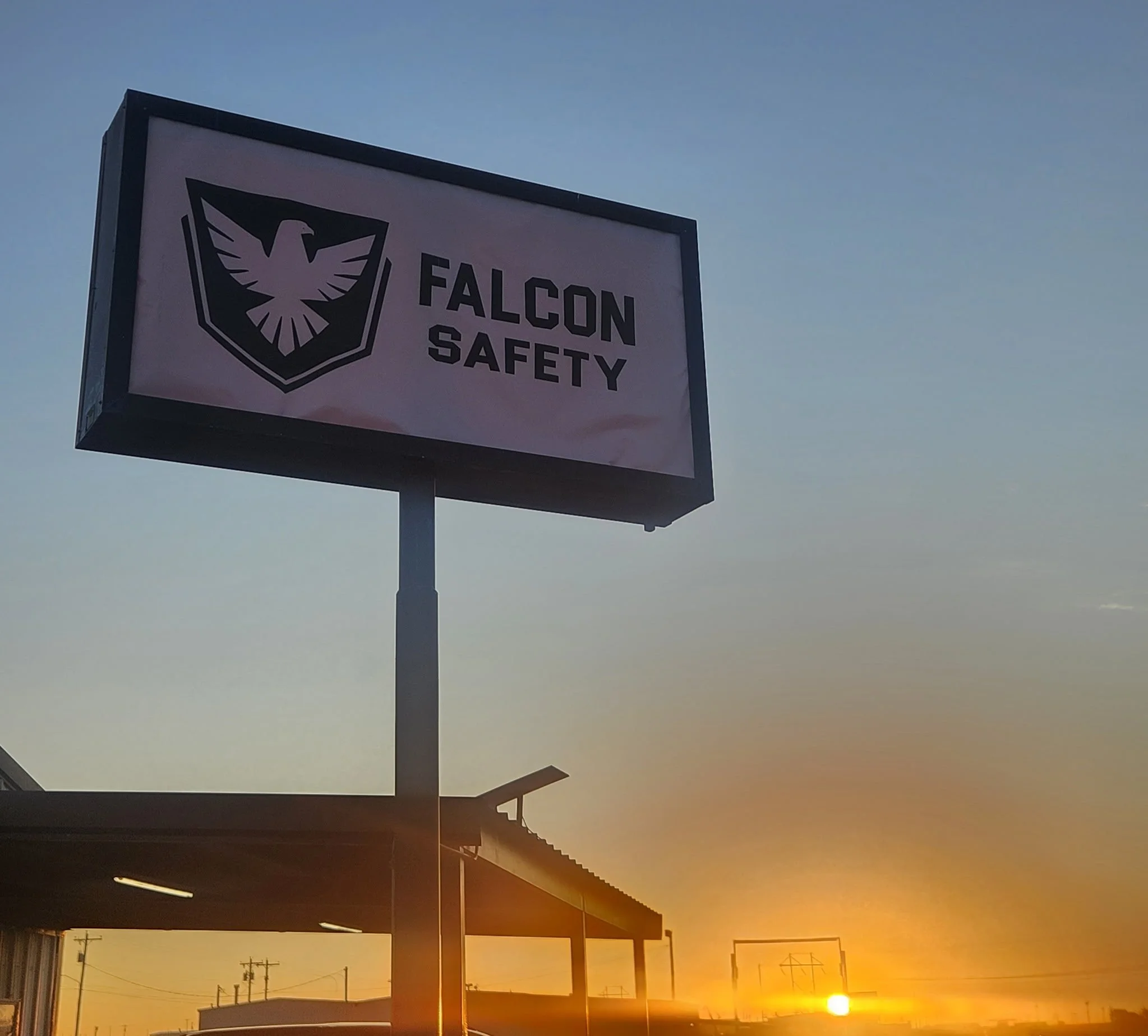 A sign reading 'Falcon Safety' beneath a geometric falcon emblem, with a sunset sky in the background.