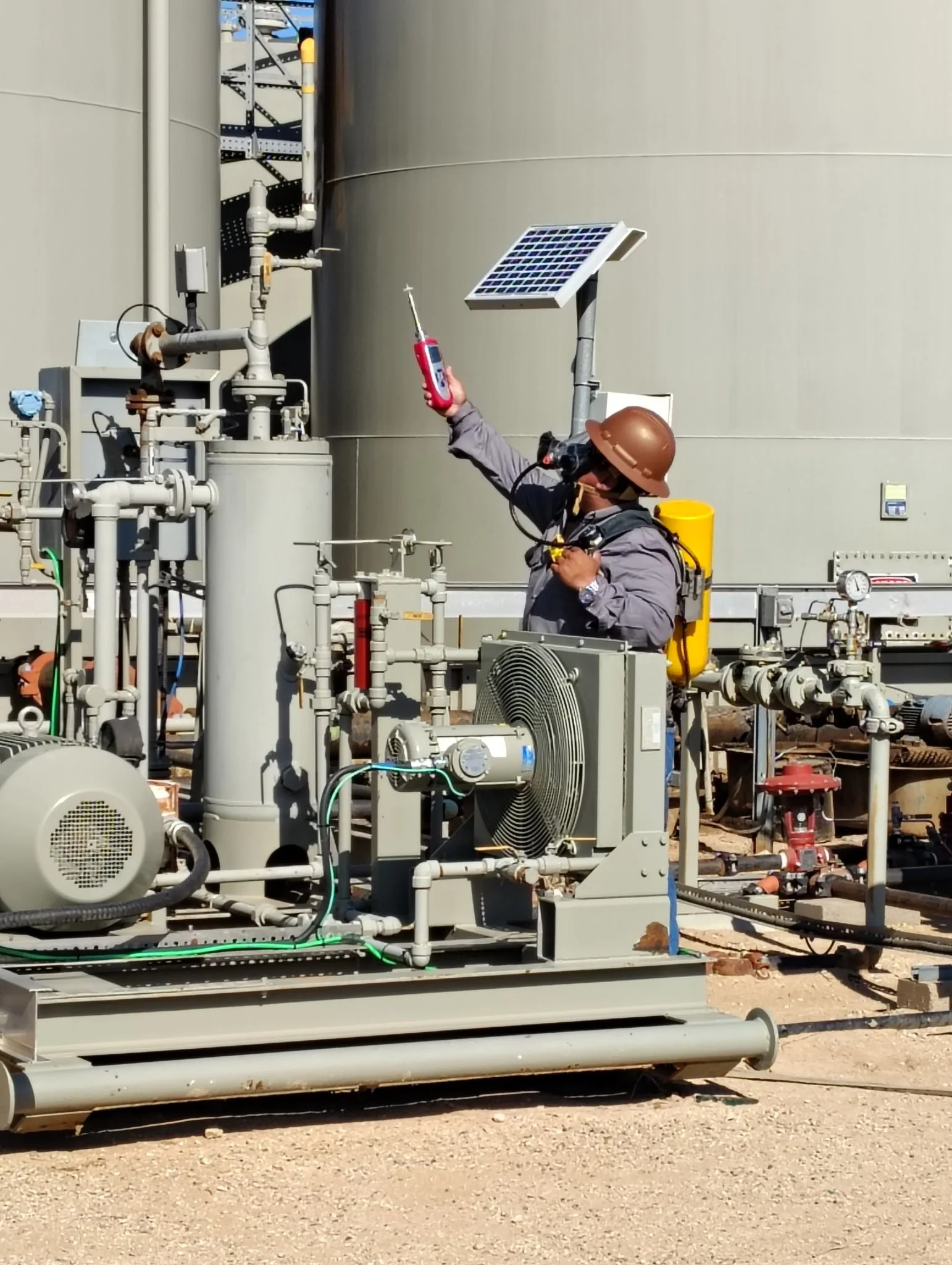 Industrial equipment with pipes, a small solar panel, and a worker in safety gear holding a digital device, situated outdoors on a dirt surface.