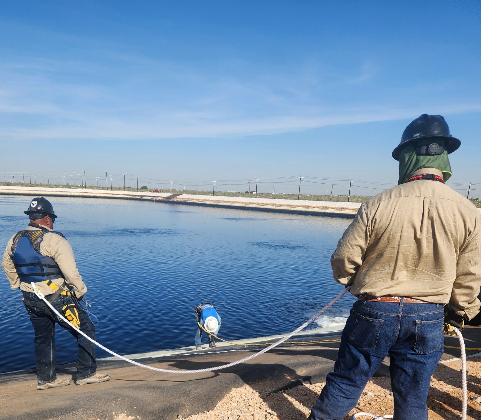 People standing on the edge of a body of water, wearing helmets and safety gear, possibly involved in construction or inspection work, with power lines in the background and a clear blue sky.