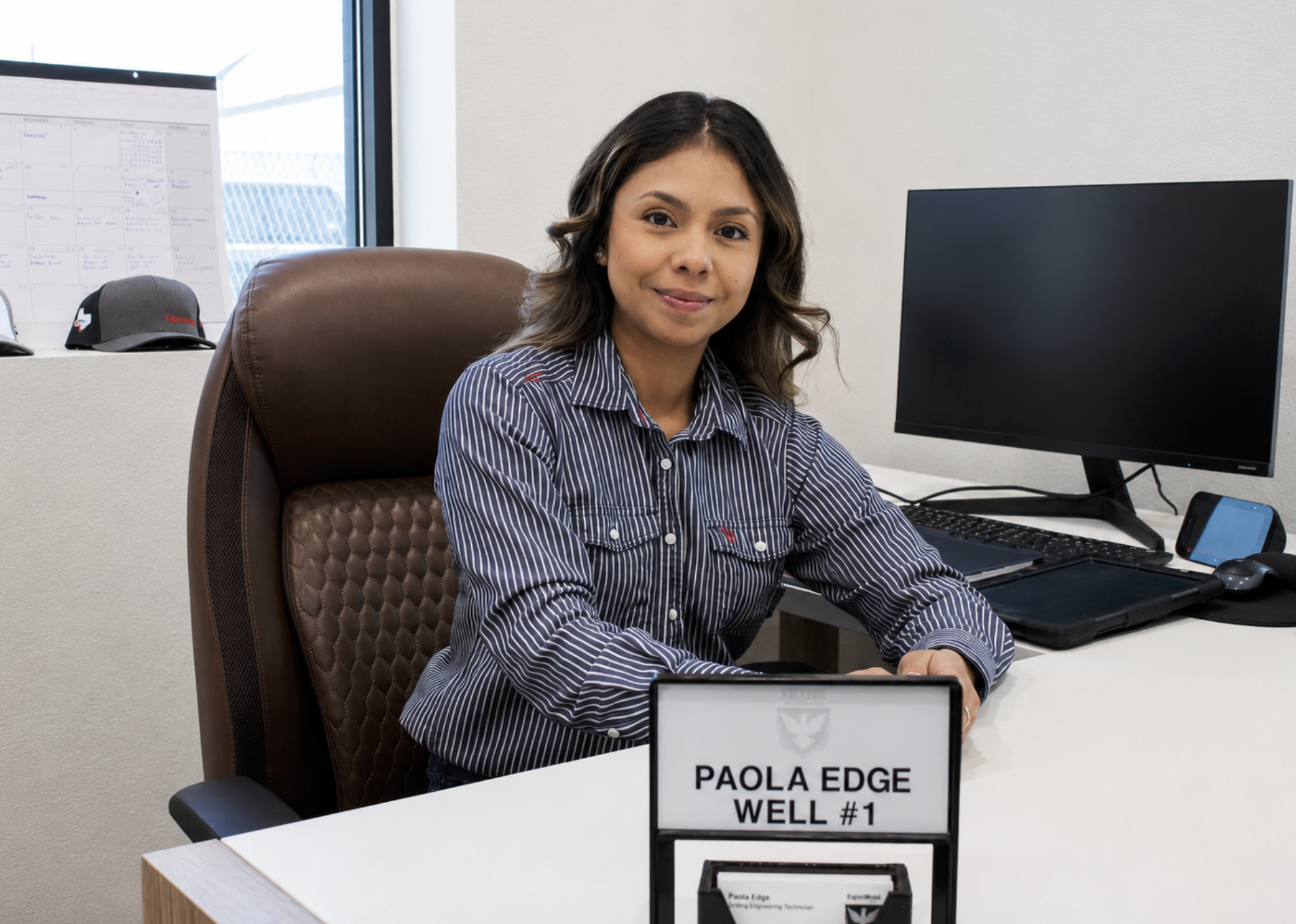 A woman sitting at a desk in an office, smiling, with a computer monitor and a nameplate that reads 'PAOLA EDGE WELL #1' in front of her.