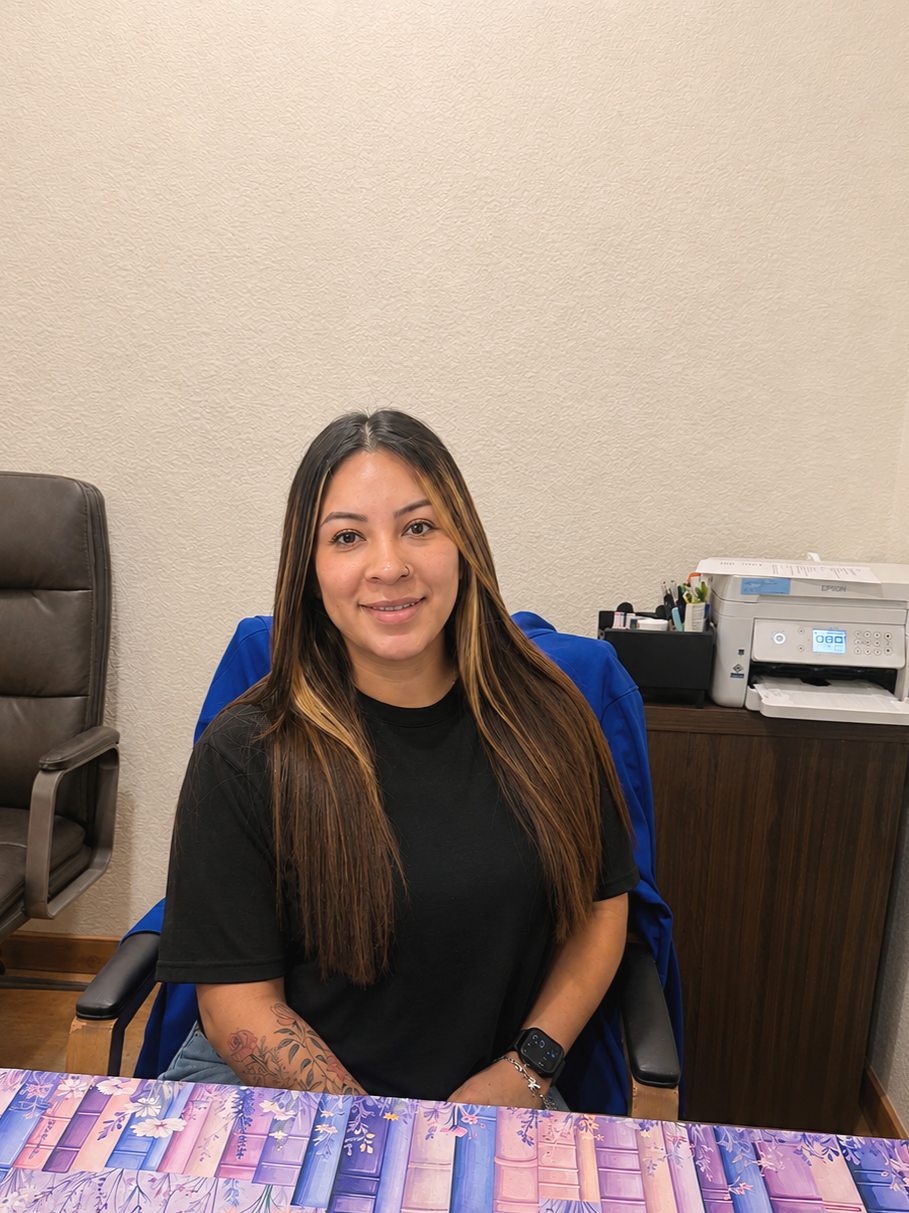 A woman with long brown hair, wearing a black t-shirt, sitting at a desk with a purple patterned tablecloth, smiling at the camera. Behind her, a desktop printer and office supplies are visible.