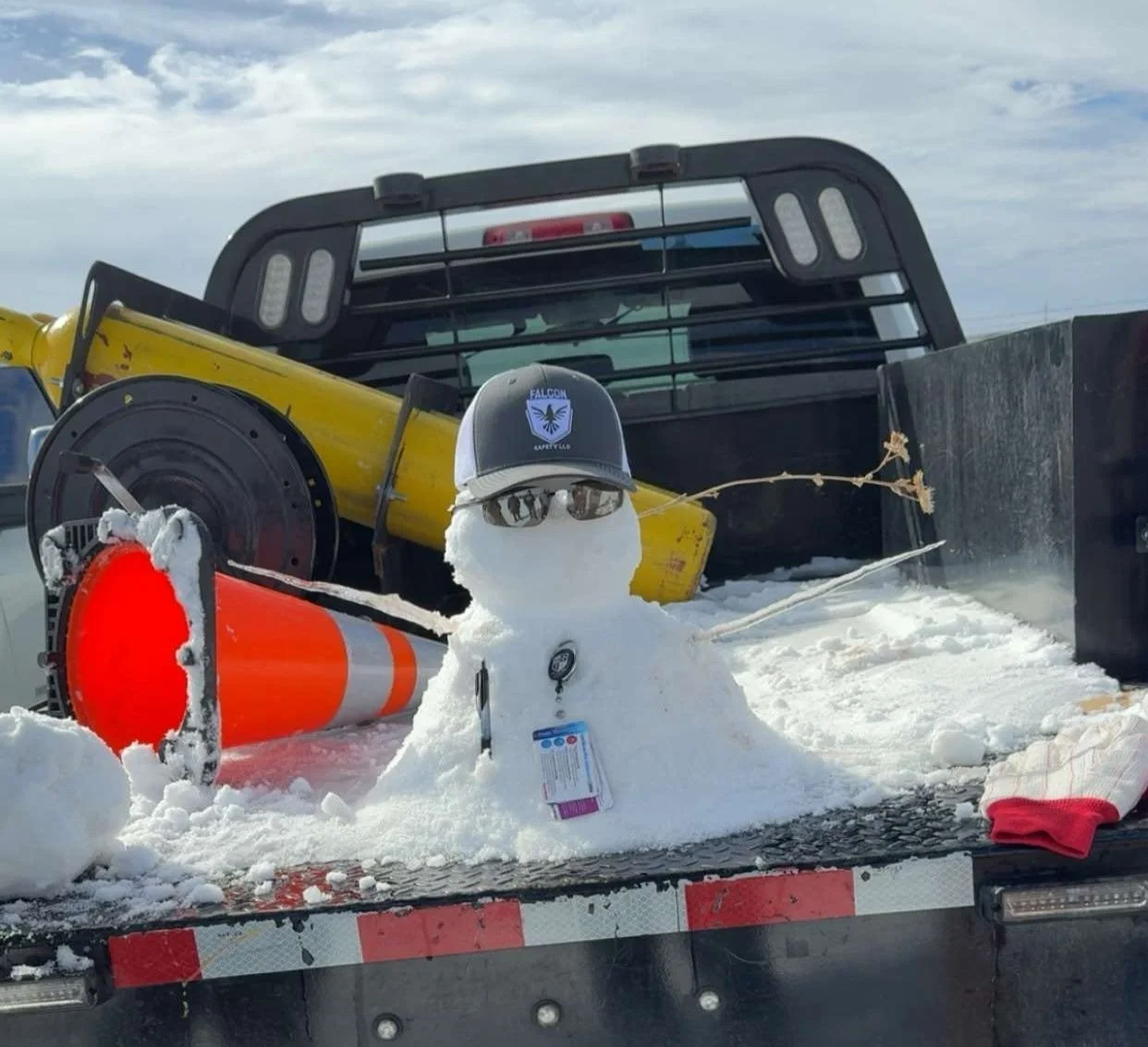 Snowman wearing sunglasses and a cap with a badge, sitting on a truck bed, surrounded by snow, an orange traffic cone, and various equipment including a yellow payload and black box, with a cloudy sky in the background.