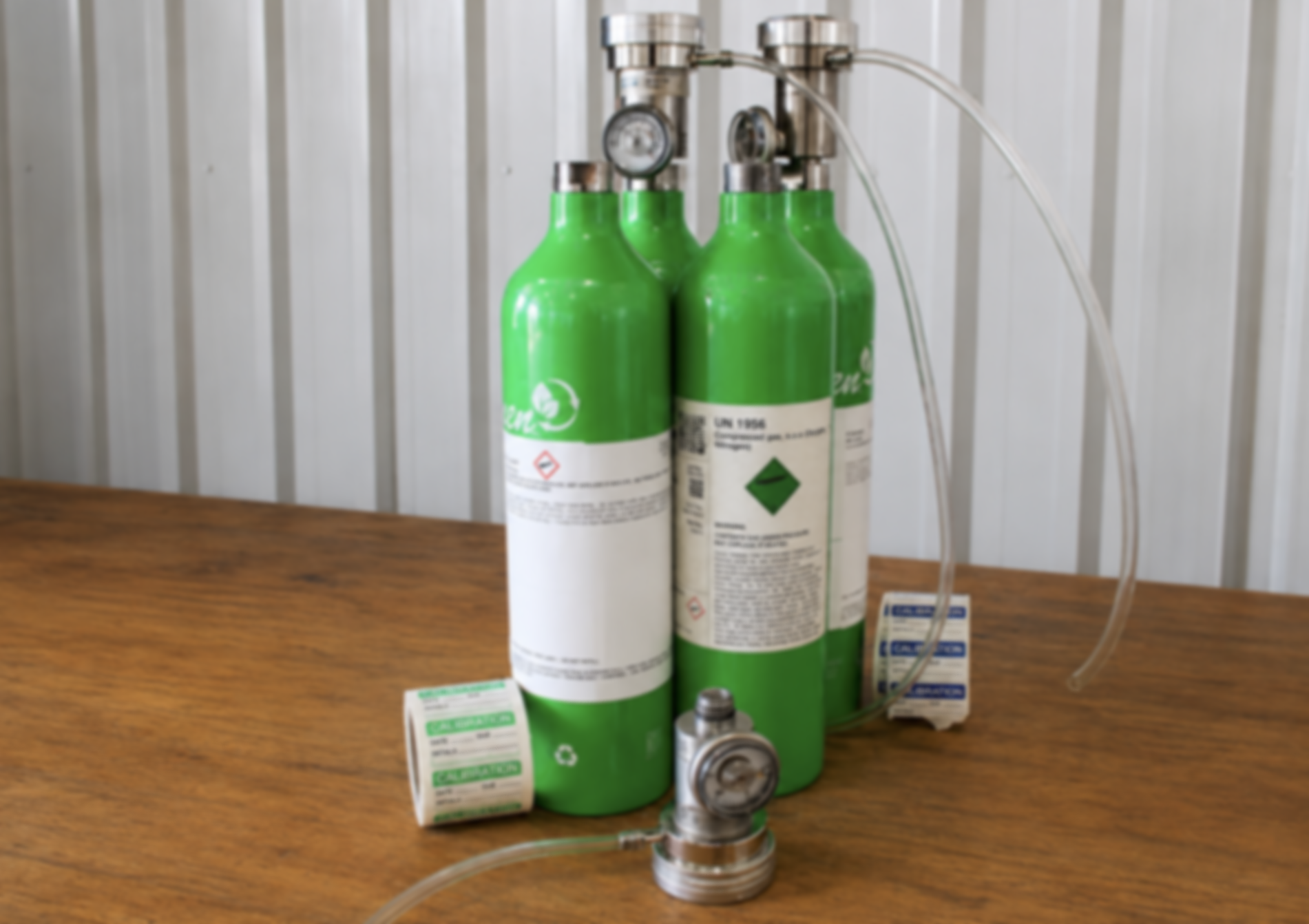 Set of green medical oxygen tanks connected to a regulator and flow meter with tubes, placed on a wooden surface against a white wall.