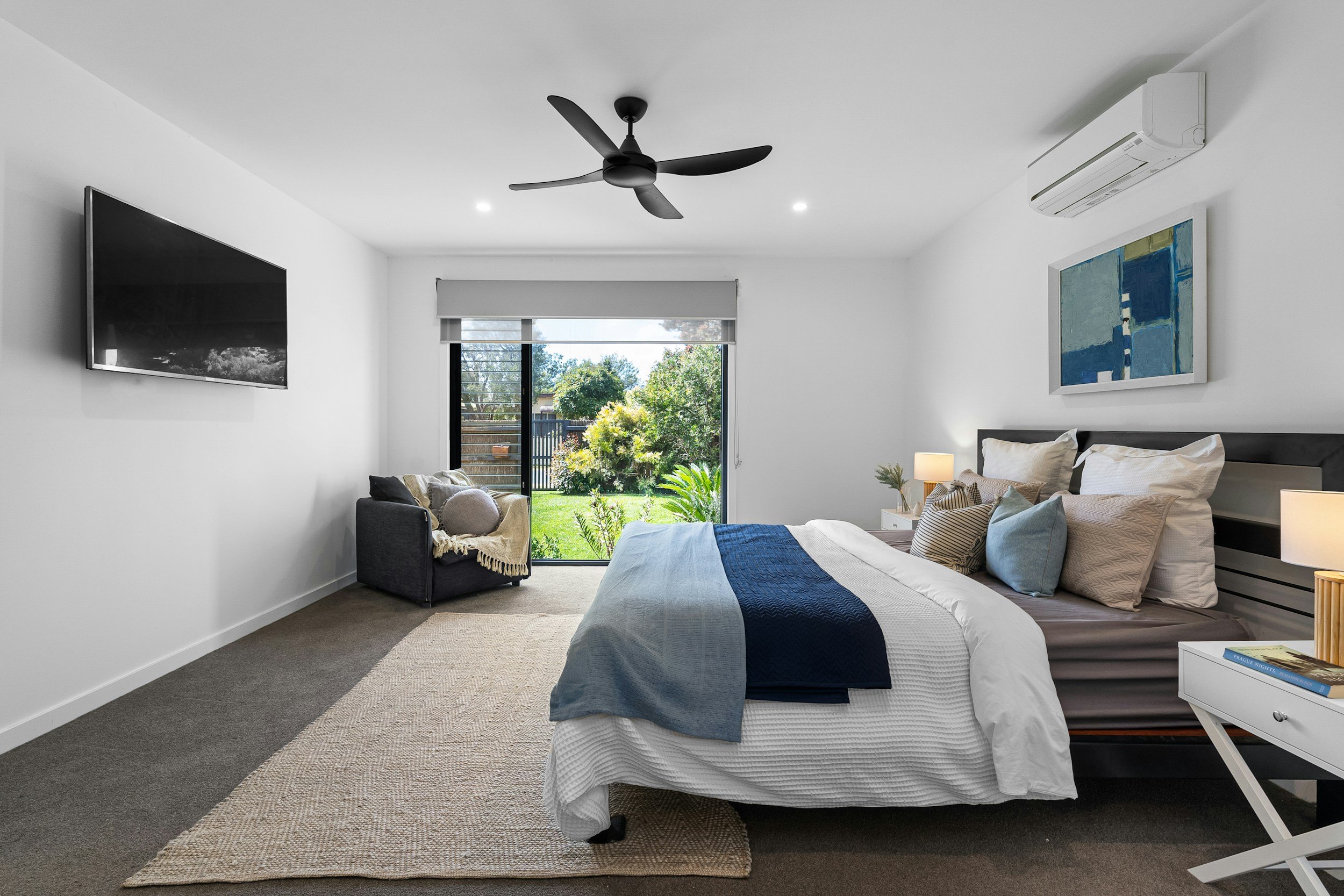 A modern bedroom with a bed, pillows, and blankets, a side table with a lamp, a chair with cushions, a wall-mounted television, a large window with a view of trees and a lawn, an artwork on the wall, a ceiling fan, and an air conditioner.