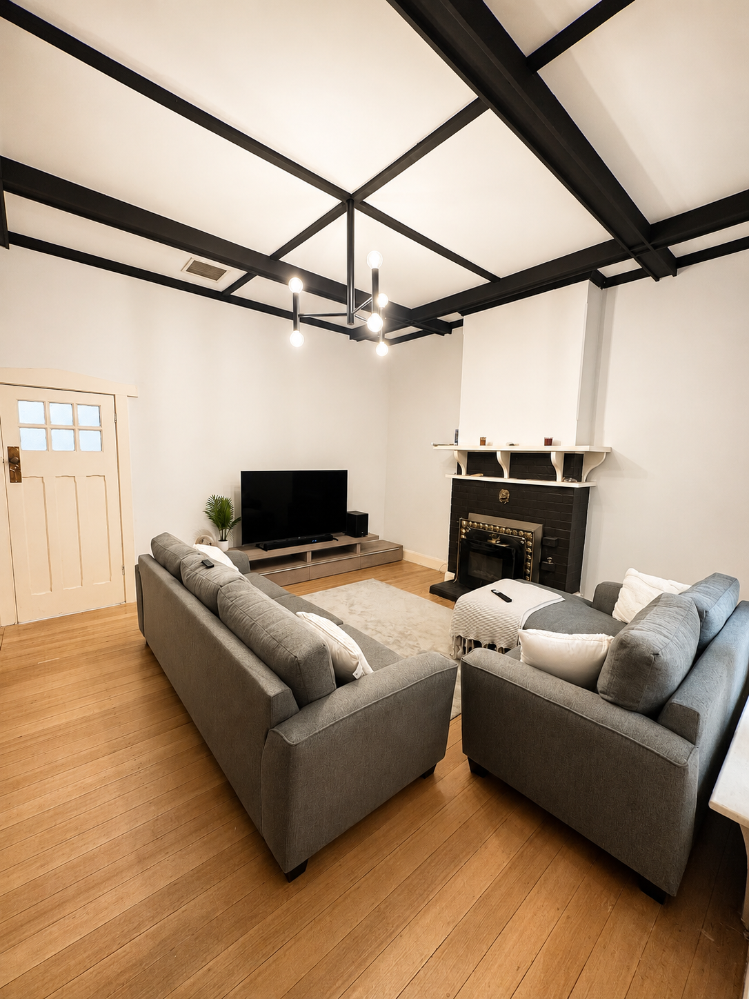 Living room with two gray sofas, a flat-screen TV on a low wooden stand, a fireplace with a black brick surround and white mantel, and hardwood floors.