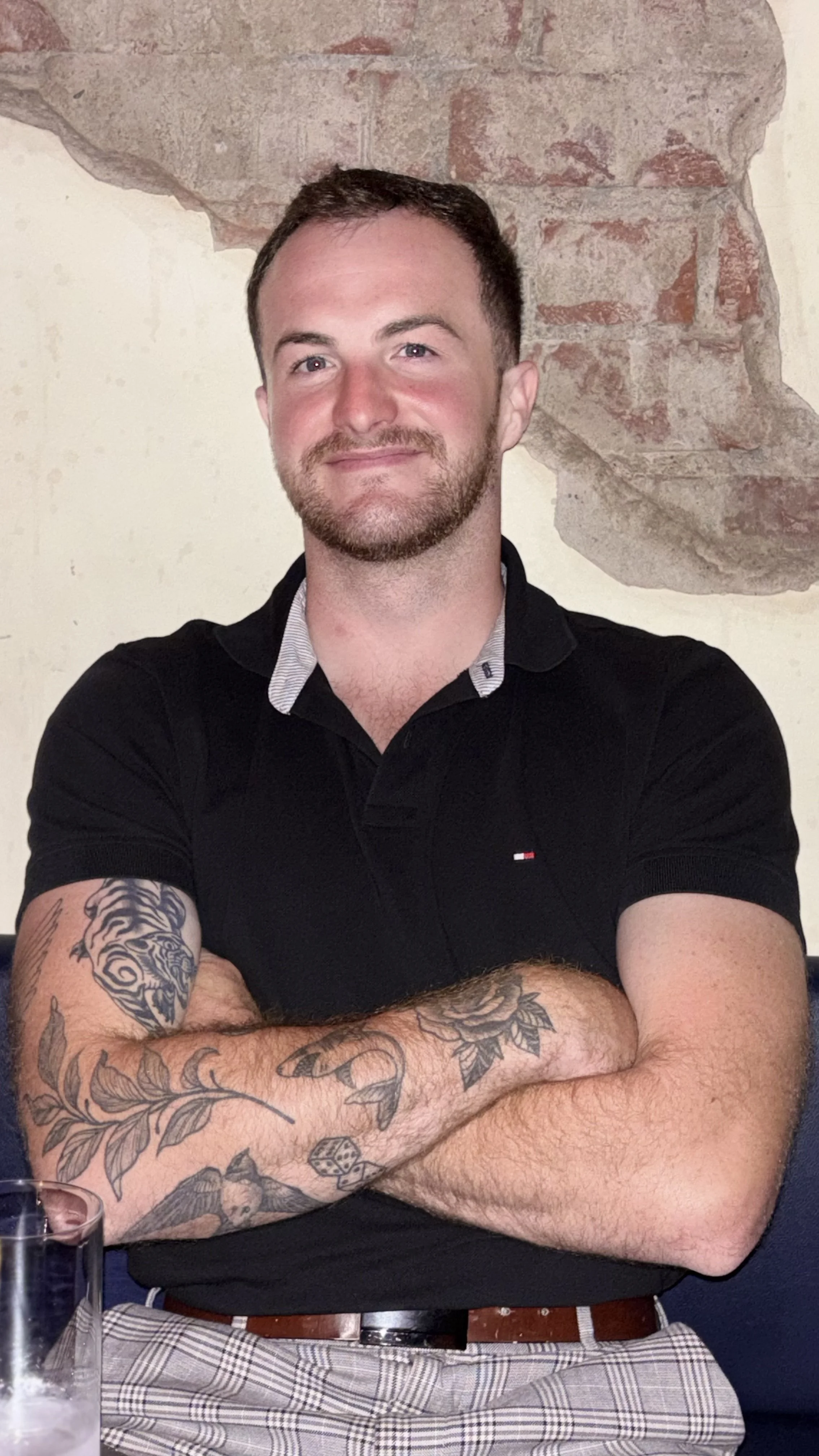 A man with short dark hair and a beard, wearing a black polo shirt, sitting with his arms crossed, in front of a brick wall. He has visible tattoos on his forearms.