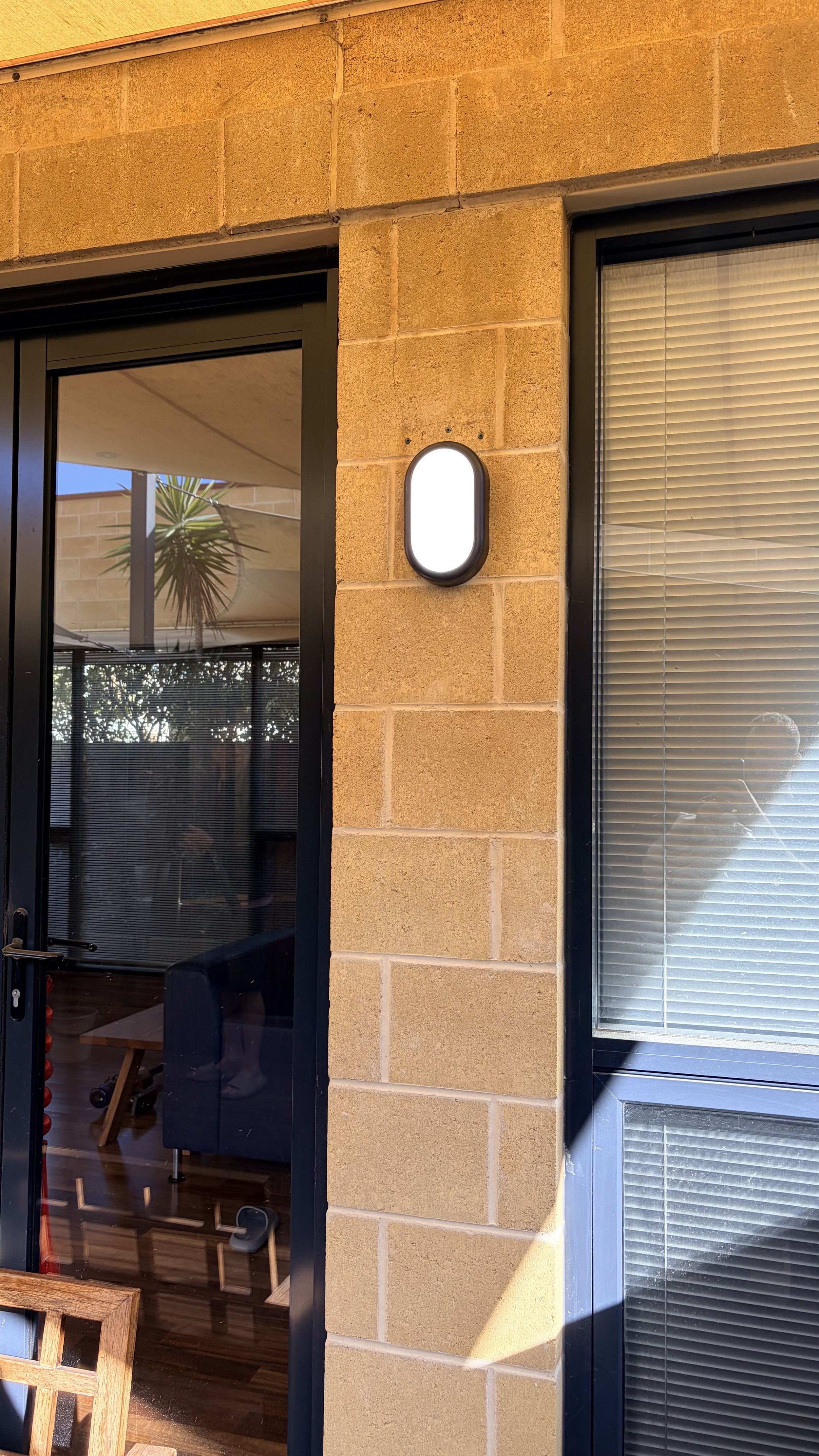 Exterior view of a building with beige brick walls, a black-framed glass door, a window with blinds, and an oval outdoor wall-mounted light fixture.