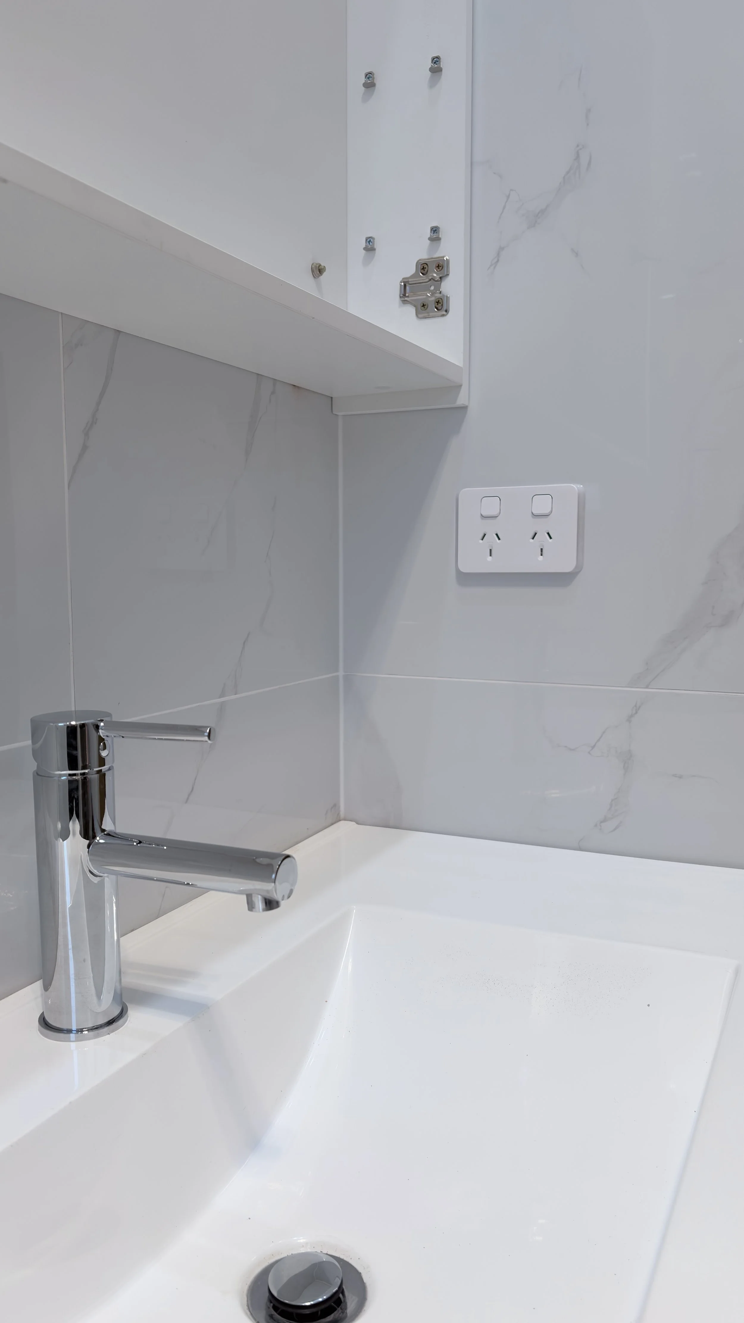 A bathroom sink with a modern chrome faucet, white countertop, and gray marble wall tiles. An electrical outlet and an unmounted cabinet door are visible on the wall.