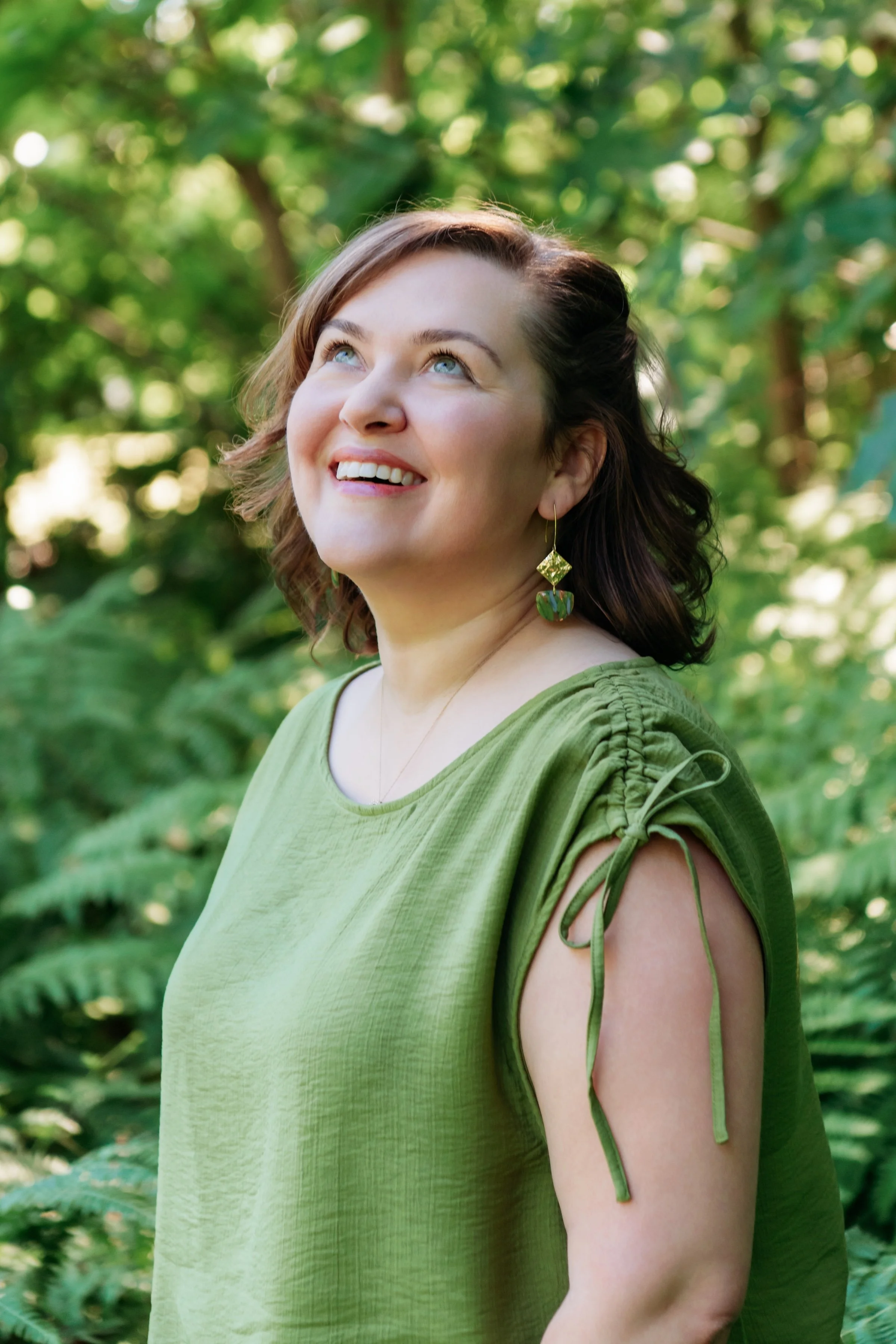 A woman with short brown hair smiling and looking upwards, wearing a green top with tied shoulder straps and earrings, standing outdoors surrounded by green foliage.
