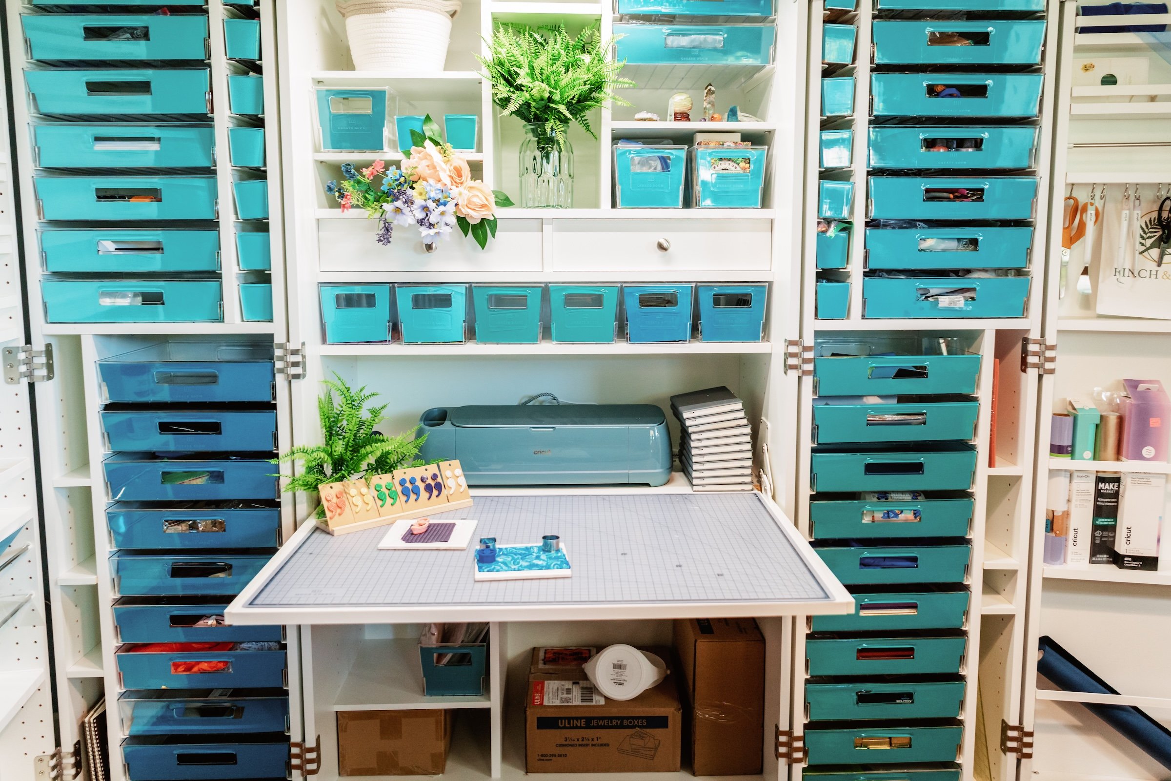 White shelving unit with multiple blue storage bins, some with transparent windows, containing craft supplies. The top of the shelf has a white basket, green fern, and a bouquet of colorful artificial flowers. The middle section has small compartments, some with contents like fabric or stickers. The bottom includes a Cricut cutting machine, a pile of papers or notebooks, and a cardboard box labeled 'Uline Jewelry Boxes.' There is a small worktable in front with crafting tools and materials.